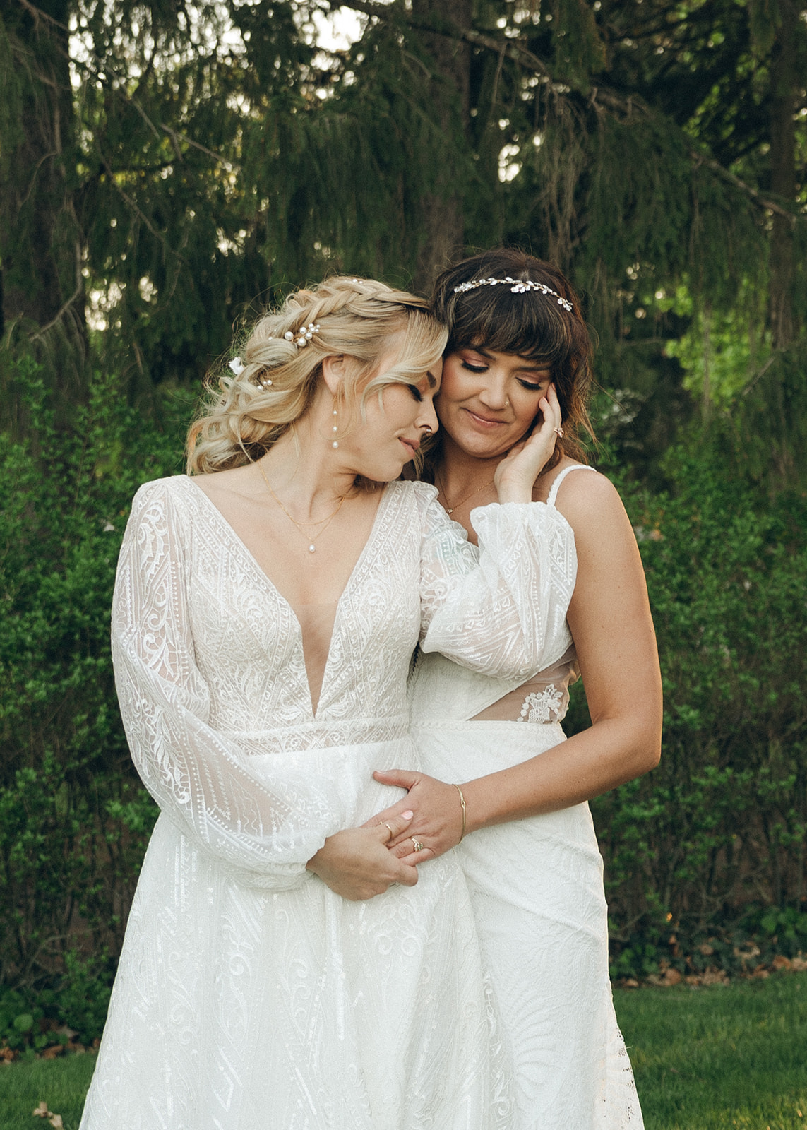 Brides in lace embroidered dresses hold each other while snuggling on a tree line