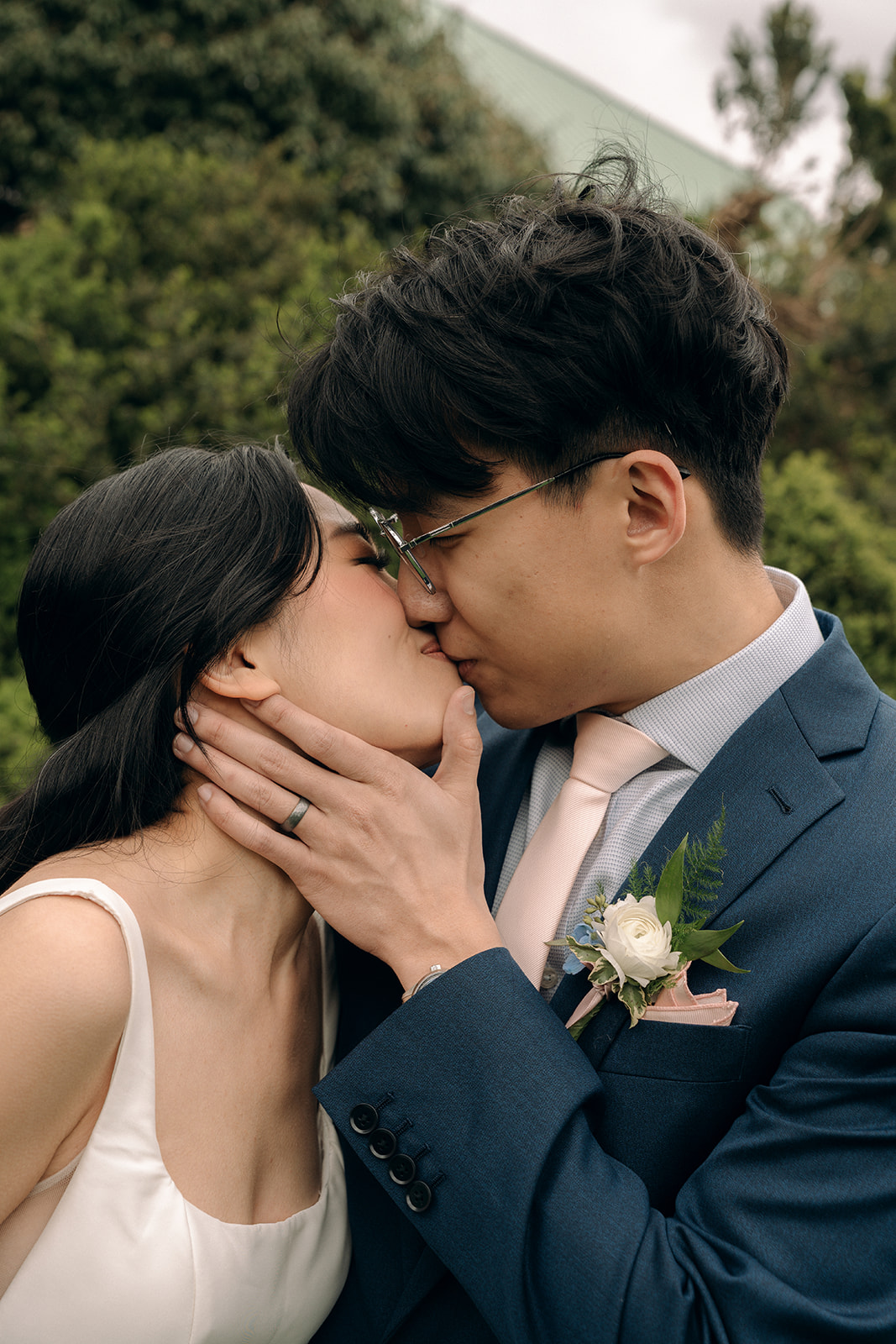 A groom kisses his bride while standing in a blue suit and peach tie