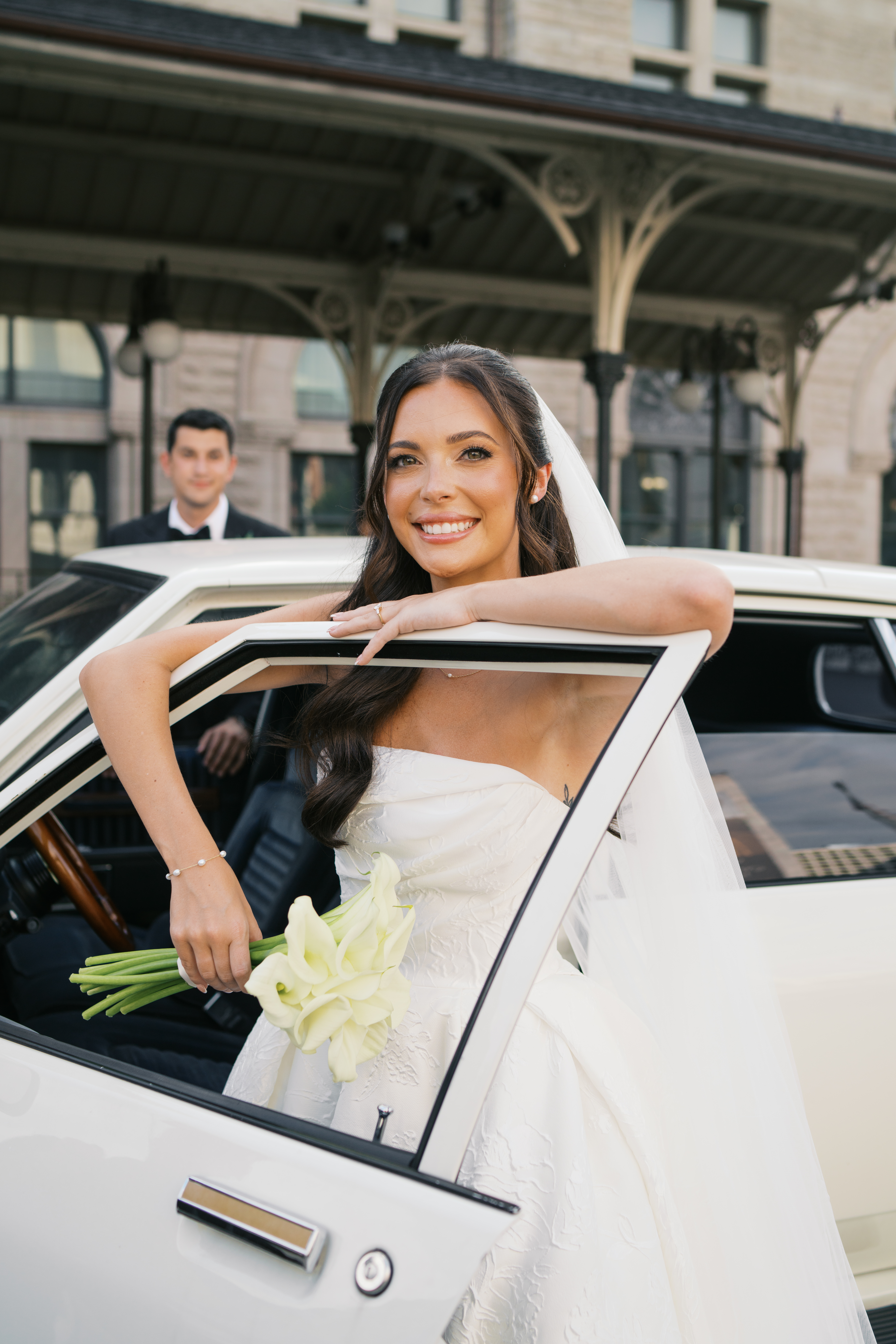 Bride with her arm up on a car window looking at the camera with the groom behind her out of focus outside of Union Station Hotel in Nashville, TN. 