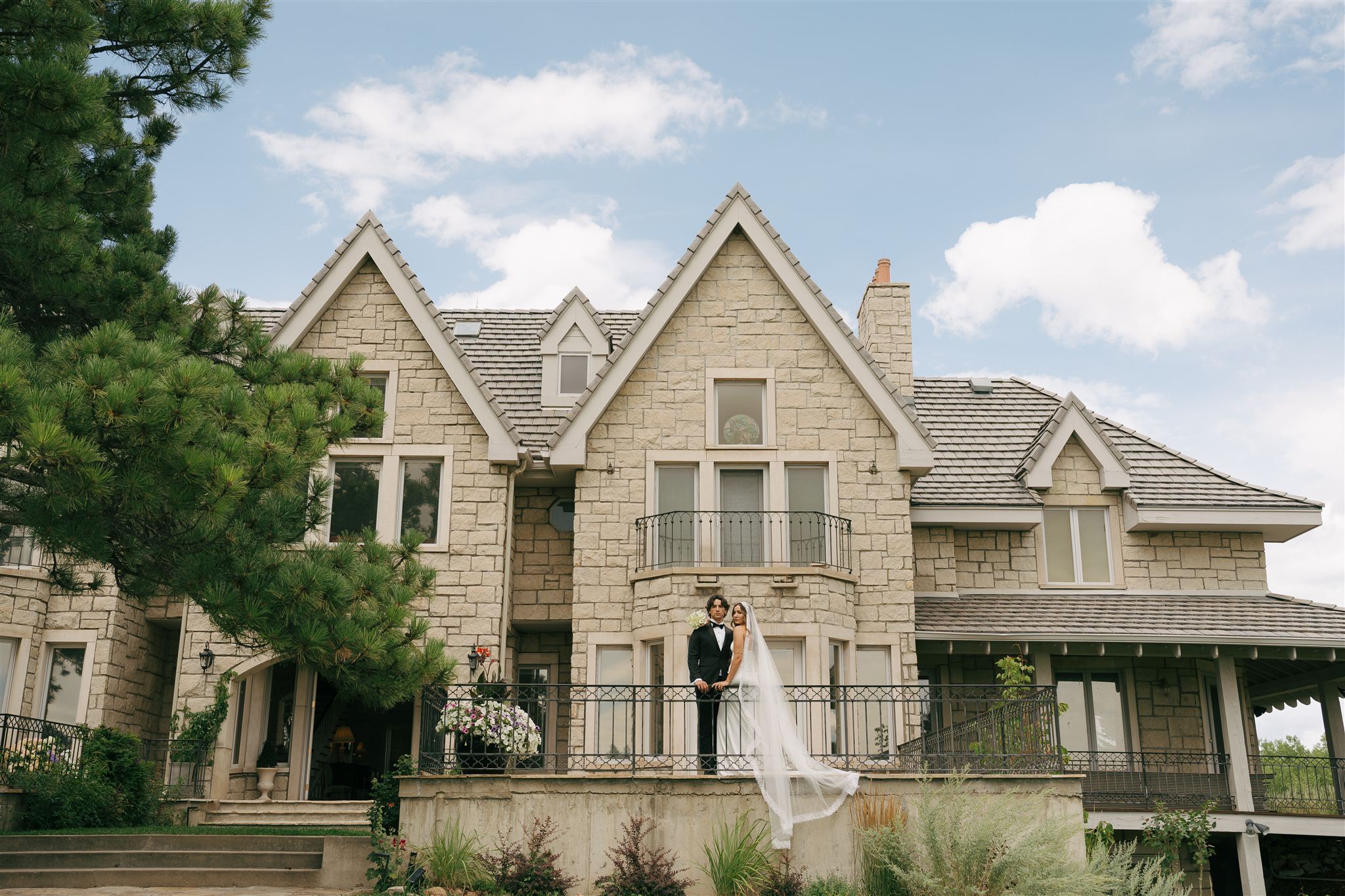 Bride and groom standing on a patio with a castle behind them with blue skies and a green tree. 