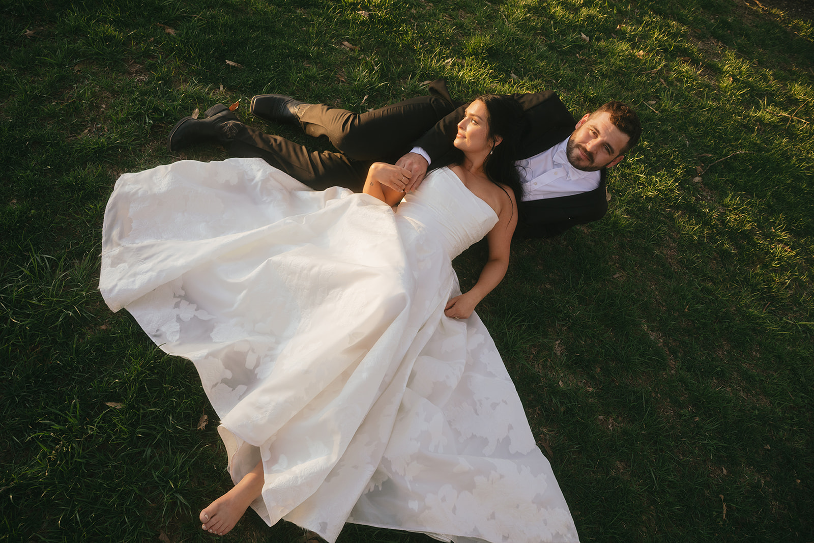 A bride lays in the lap of her groom in a green lawn at sunset during their Nashville destination wedding