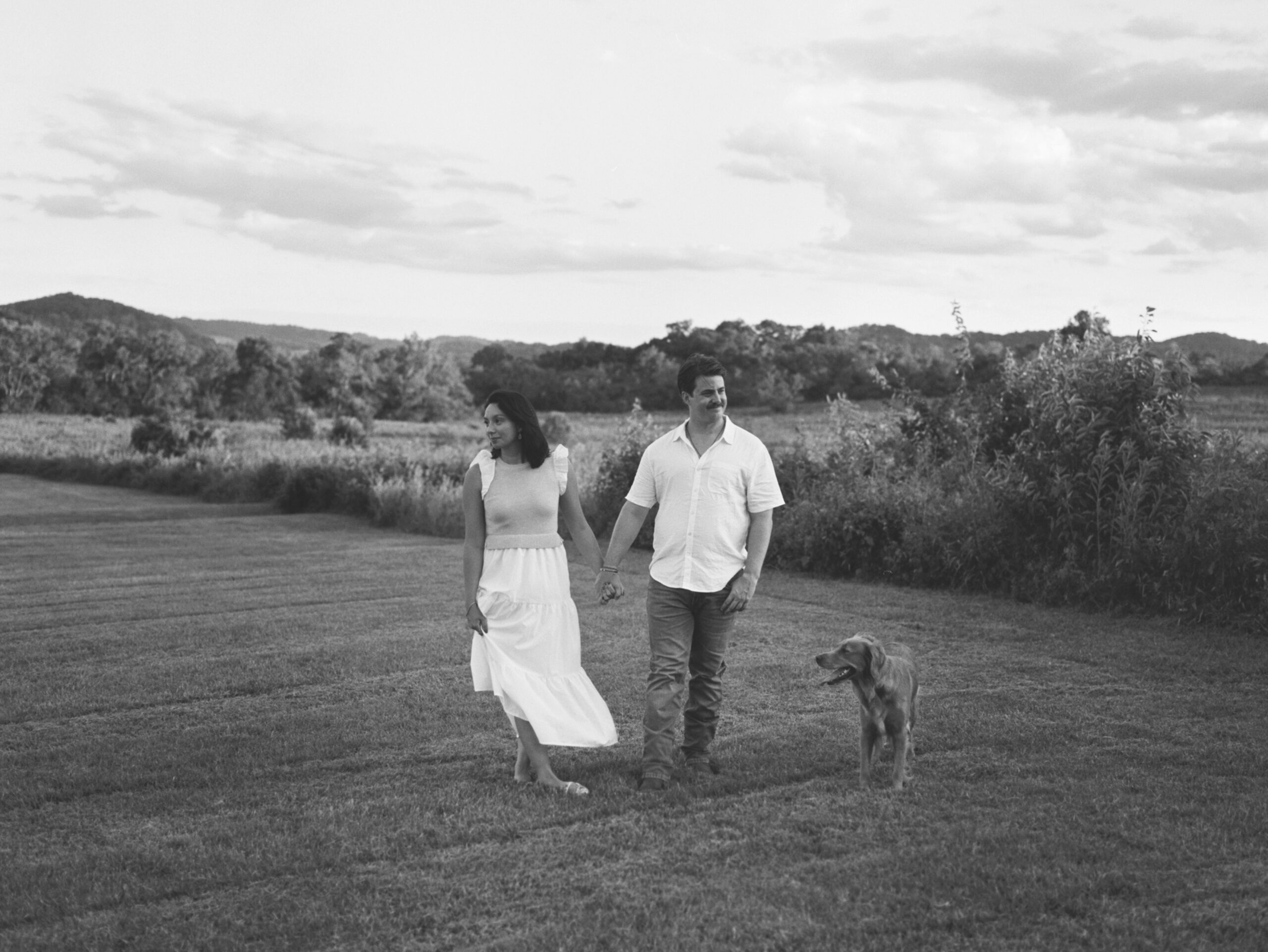 Couple standing in a field hand in hand with their golden retriever. 