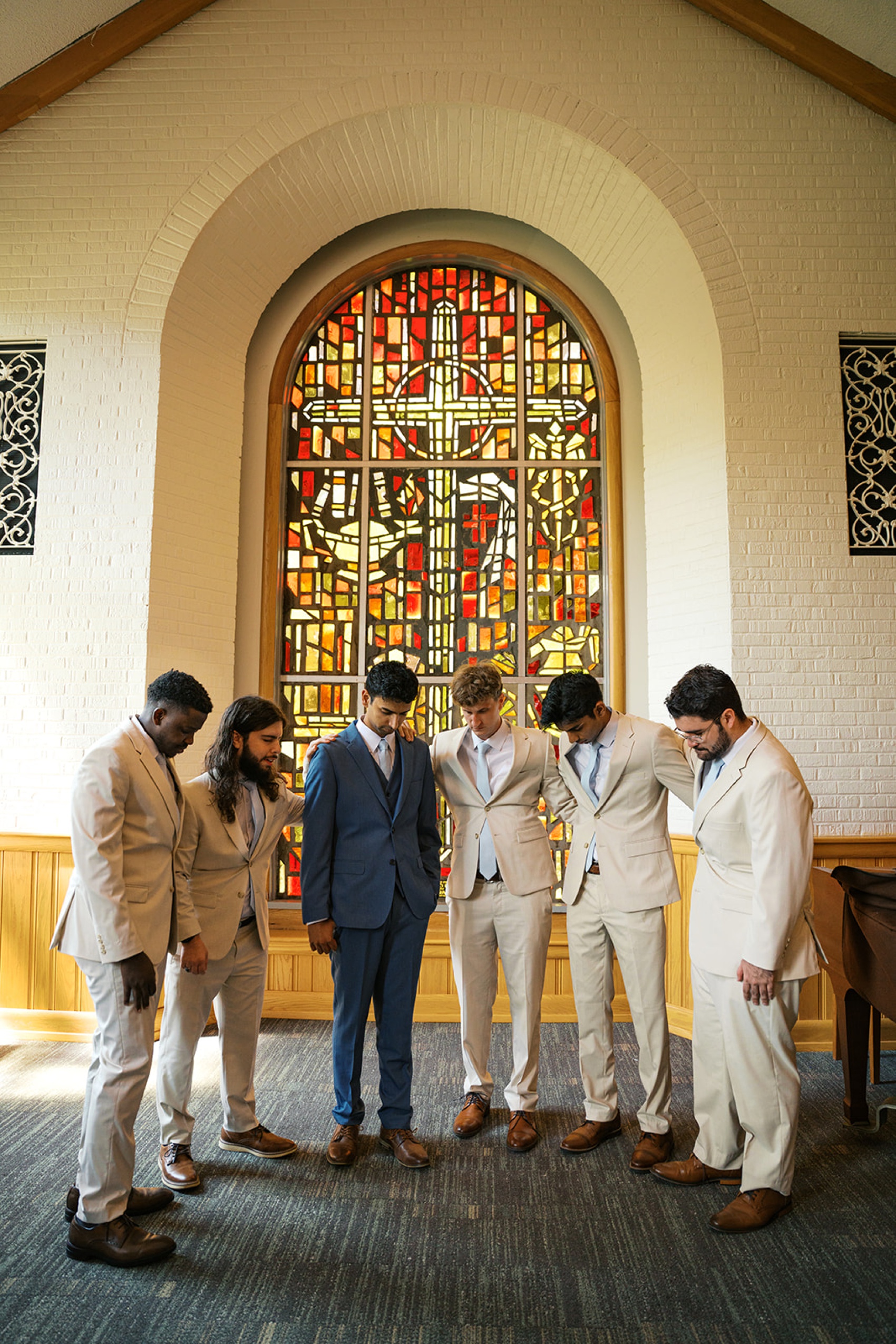 A groom prays with his groomsmen in cream suits under a stained glass window
