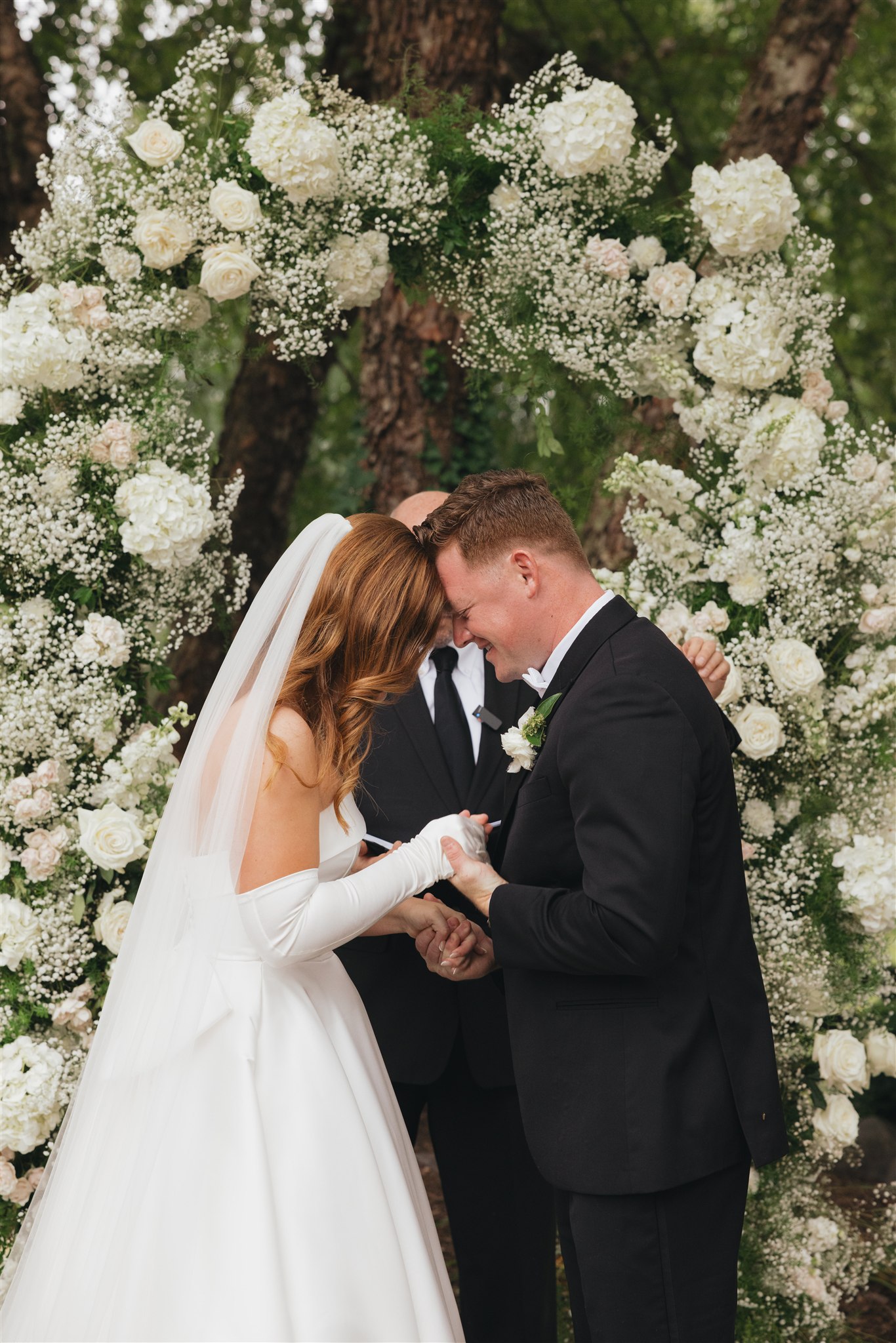 A bride and groom touch foreheads while holding hands during their outdoor ceremony under a white flower arch at one of the all inclusive wedding venues in Nashville