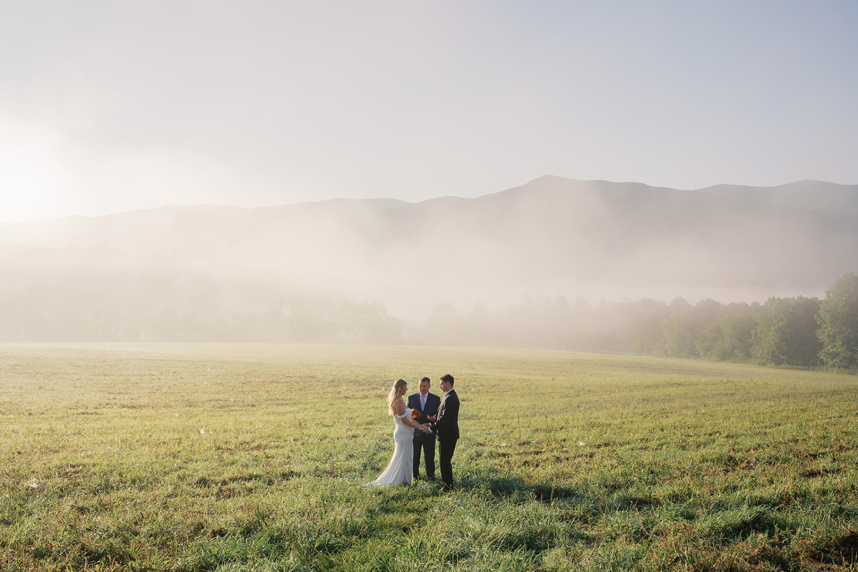 A bride and groom stand with their officiant in an open meadow covered in fog during their cades cove wedding