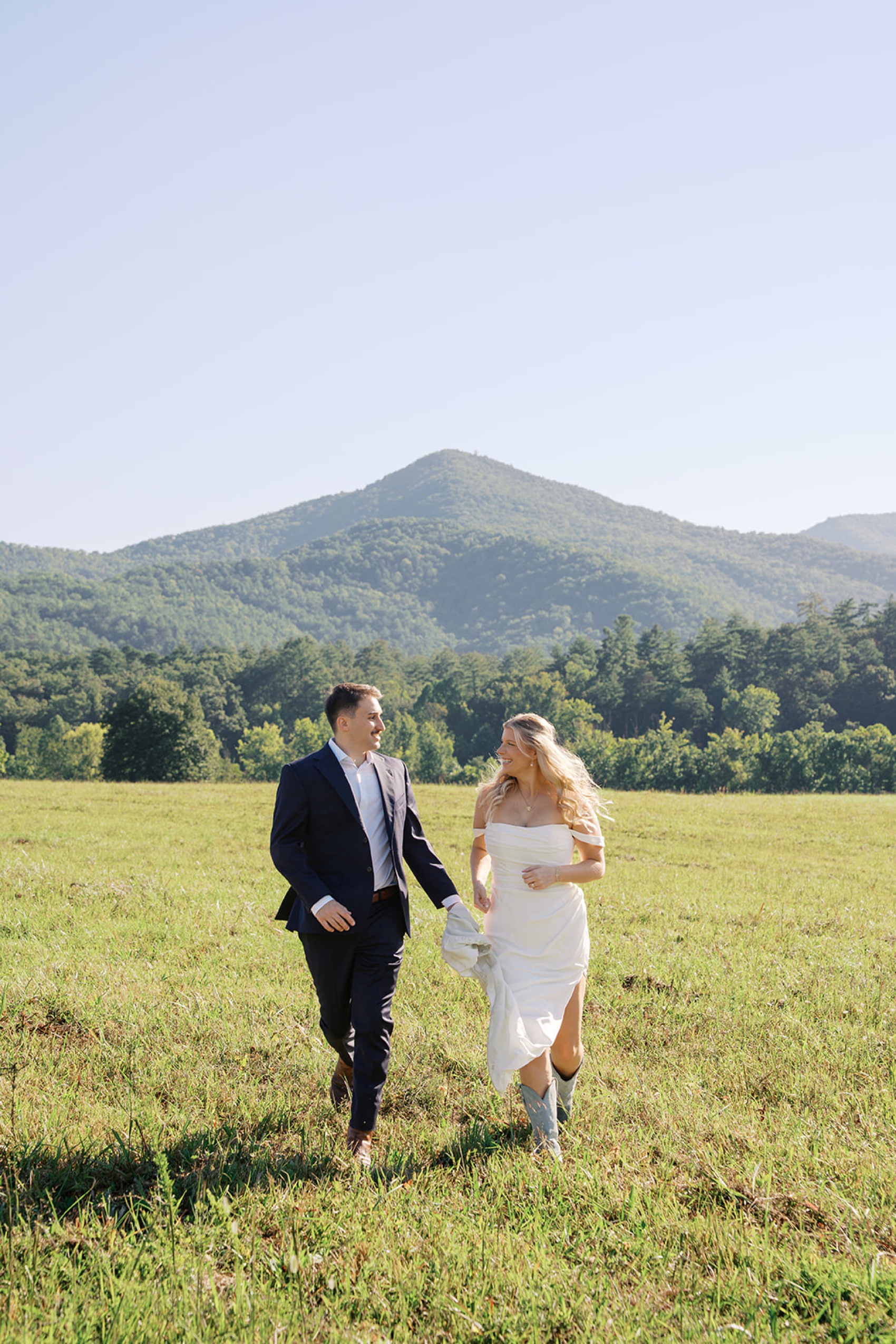 A bride and groom jog in a meadow in the mountains in boots during their cades cove wedding