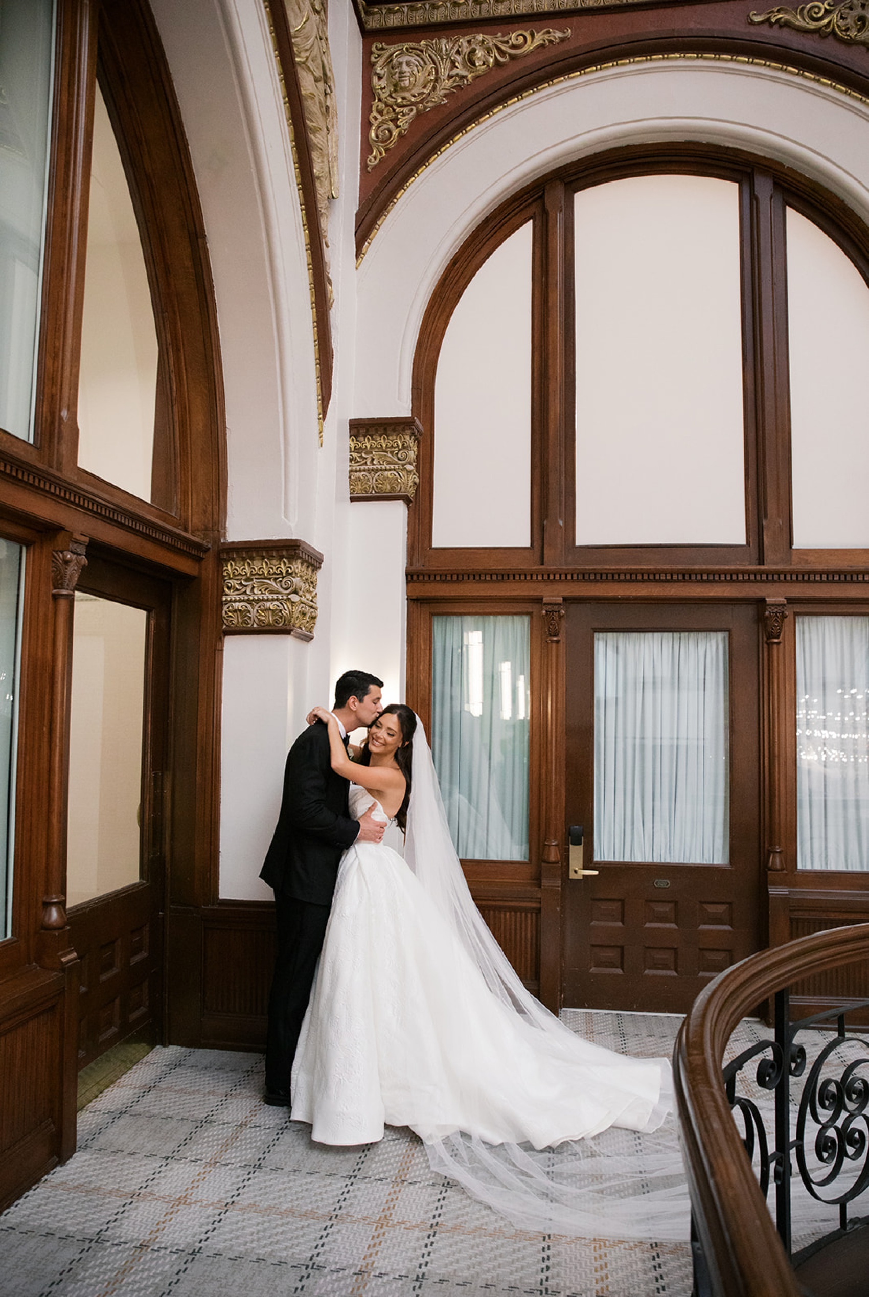 A groom kisses the temple of his laughing bride in a hallway with wood trim