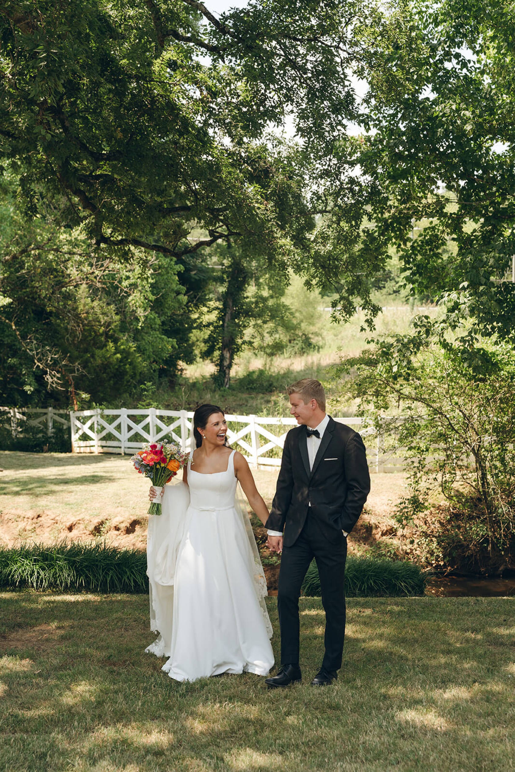 Happy newlyweds laugh while walking along a creek holding hands under tall trees