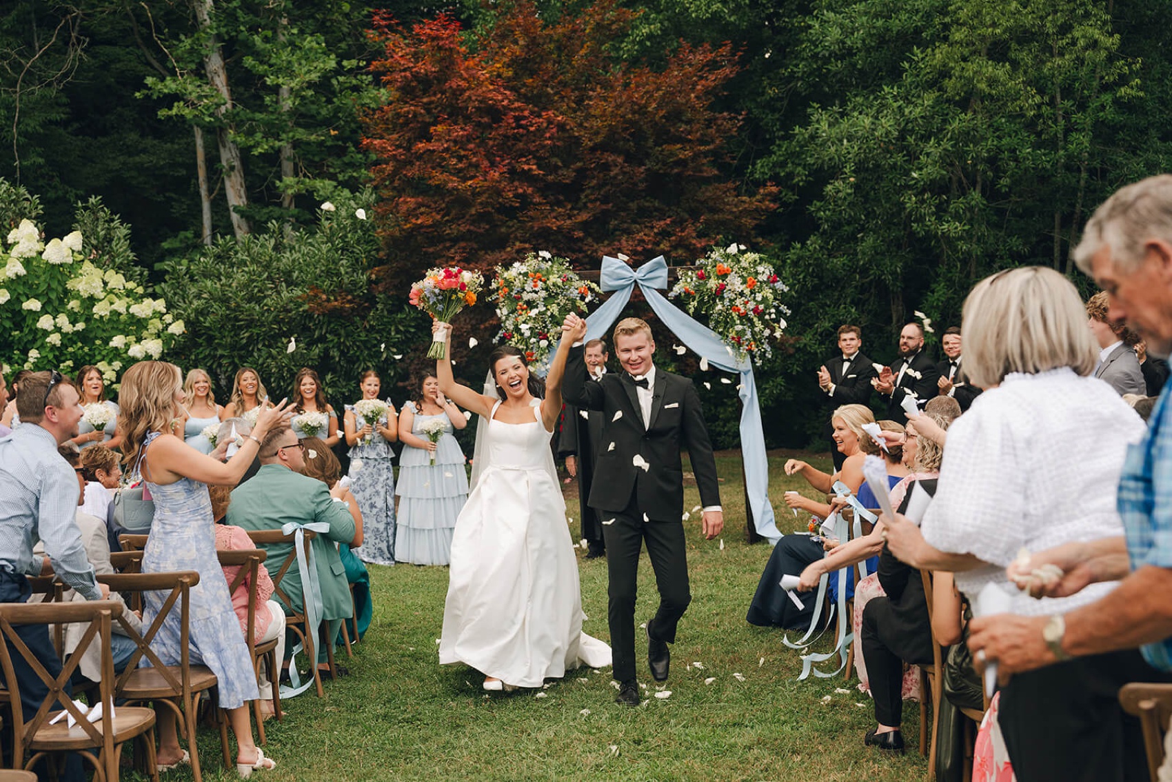 A bride and groom cheer and hold hands up while walking up the aisle from their outdoor Dara's Garden wedding ceremony as guests throw flower petals