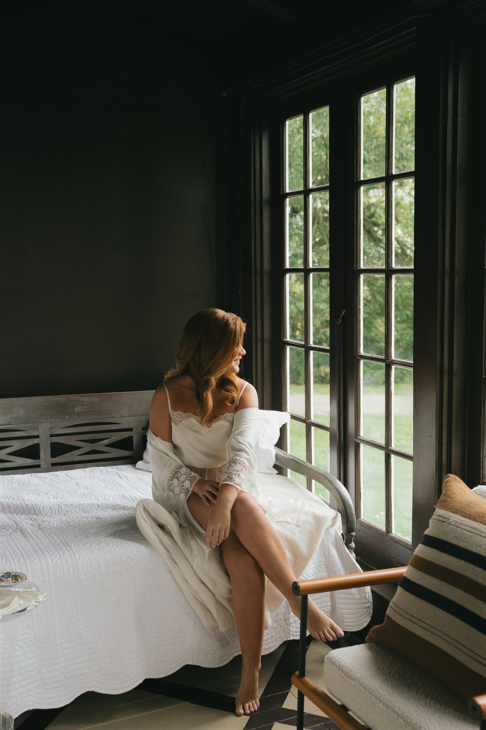 a bride sits on a bed in pajamas and looks out a window