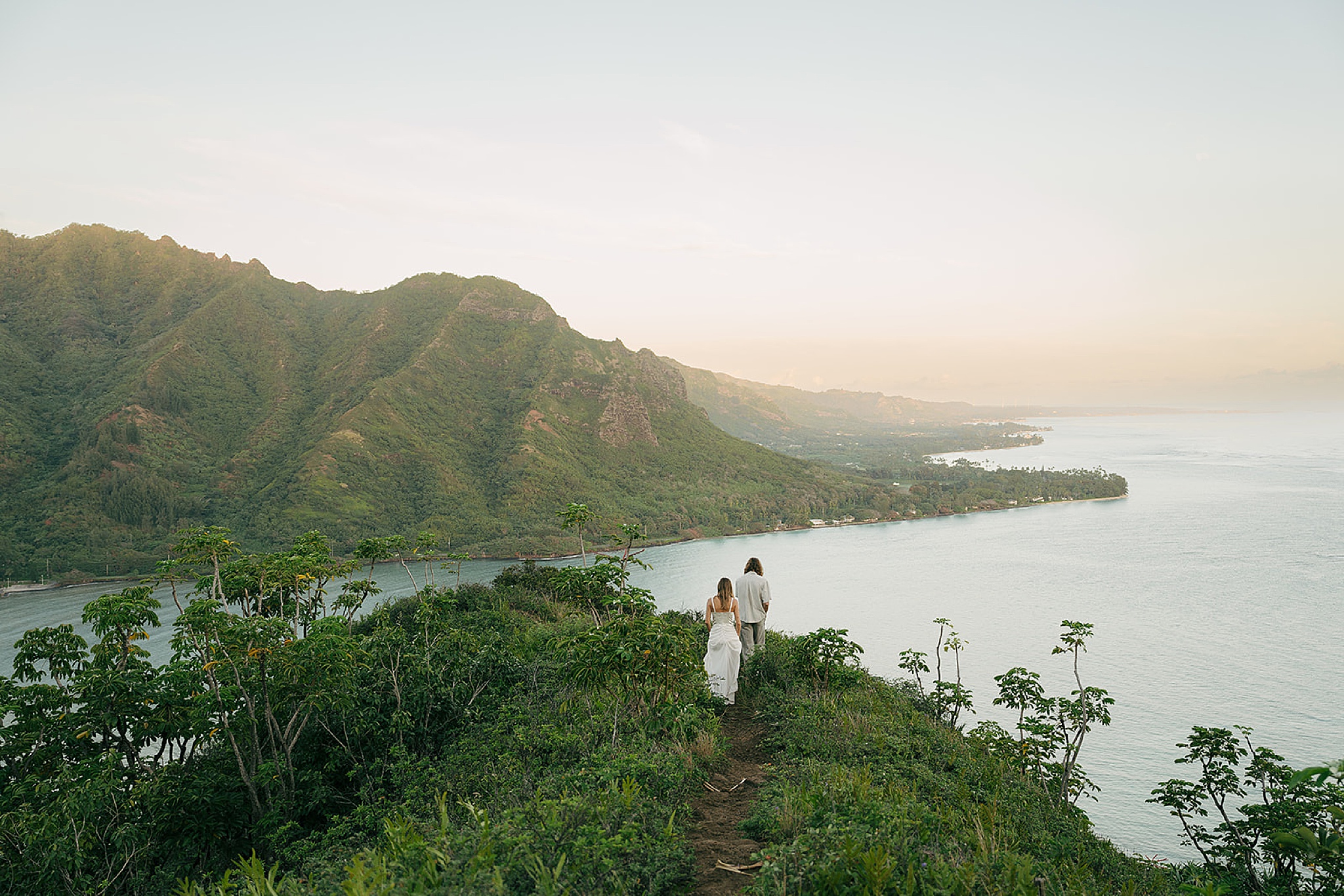 A groom leads his bride down a ridge trail overlooking the ocean at sunset