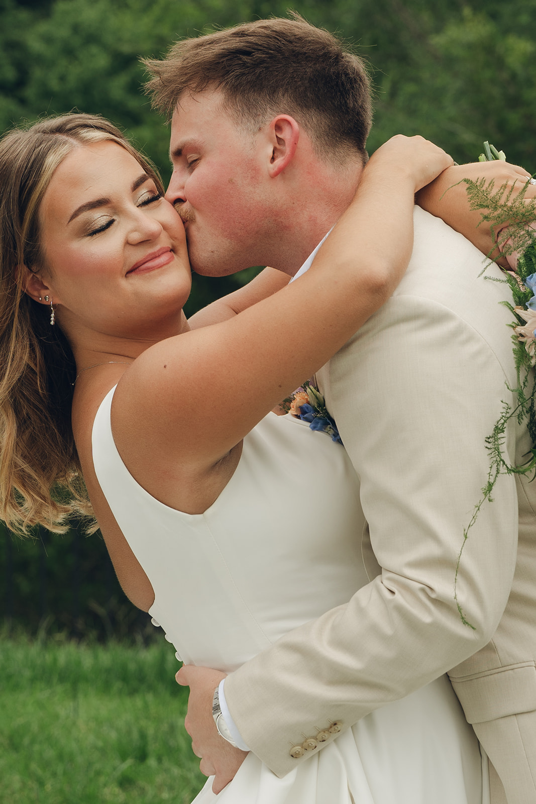 a groom in a khaki suit kisses the cheek of his smiling bride as they hug in a lawn