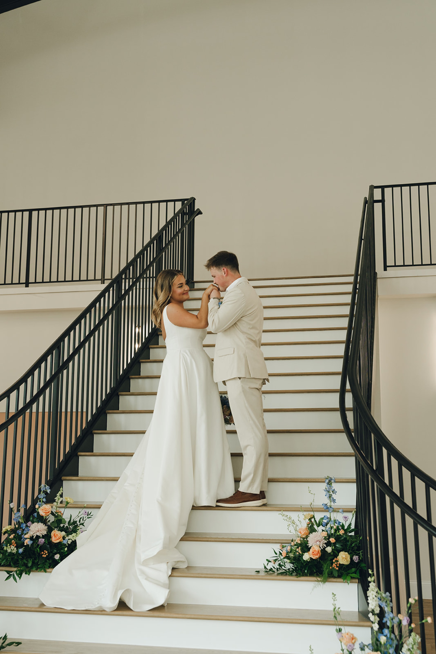 A groom in a khaki suit kisses the hand of his bride while standing on the nichols heir wedding venue grand staircase
