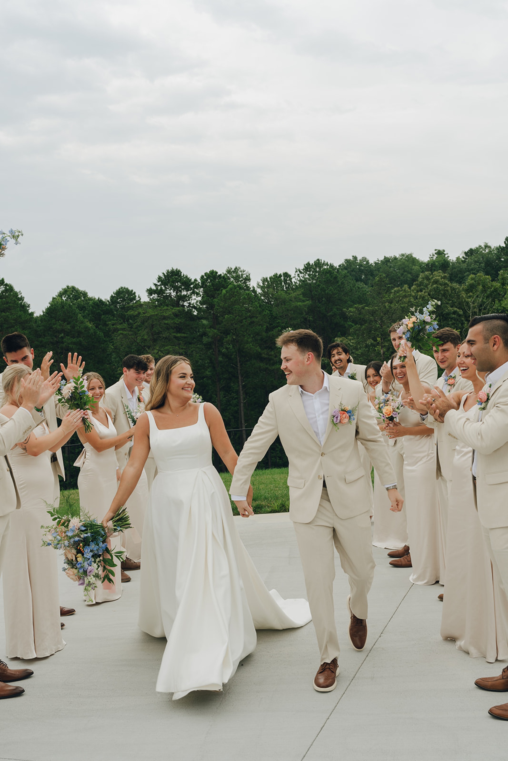 A bride and groom walk holding hands and smiling at each other during their nichols heir wedding as their wedding party cheers around them