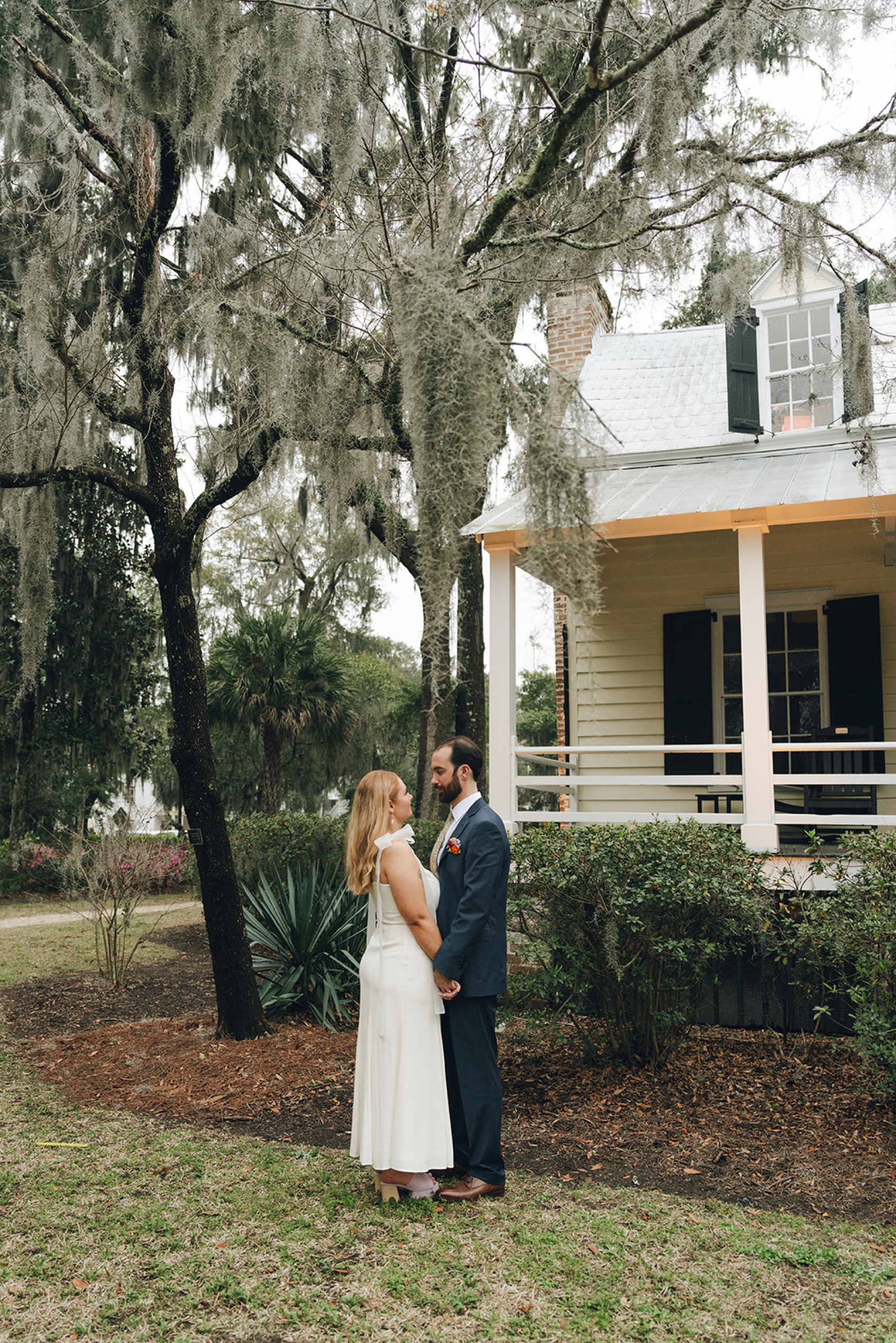 A bride and groom stand holding hands and smiling at each other in the garden by the front porch