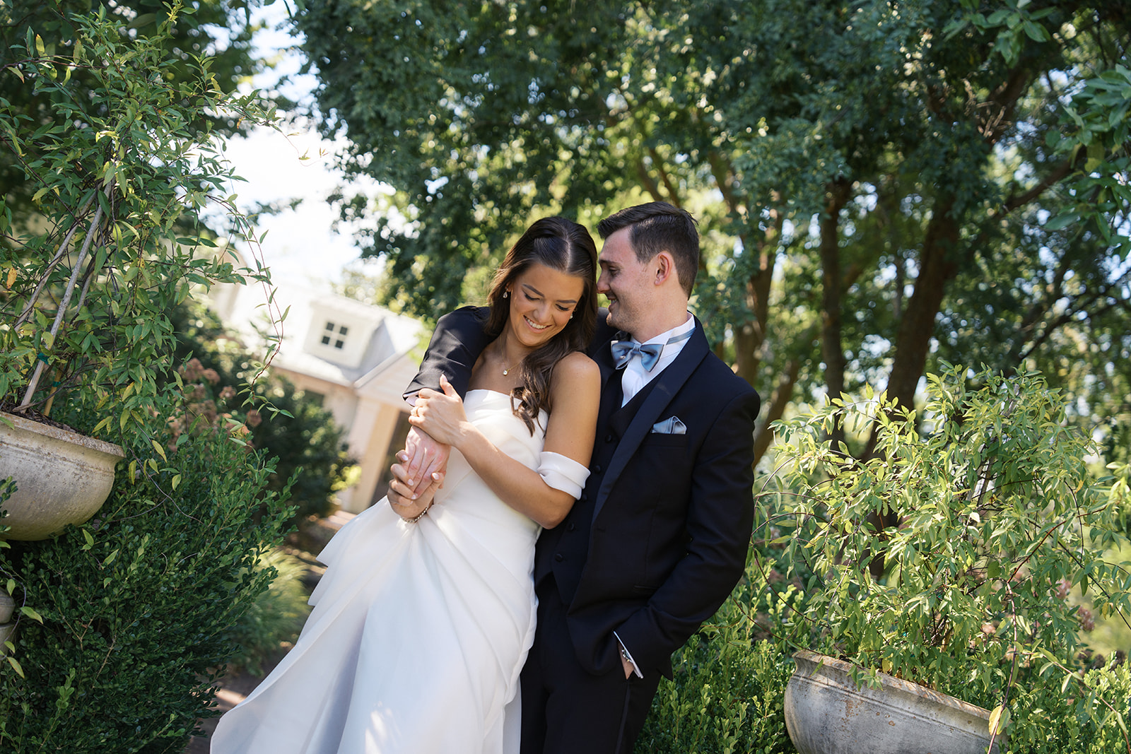 a bride hangs on the arm of her groom over her shoulder while walking in a garden and giggling