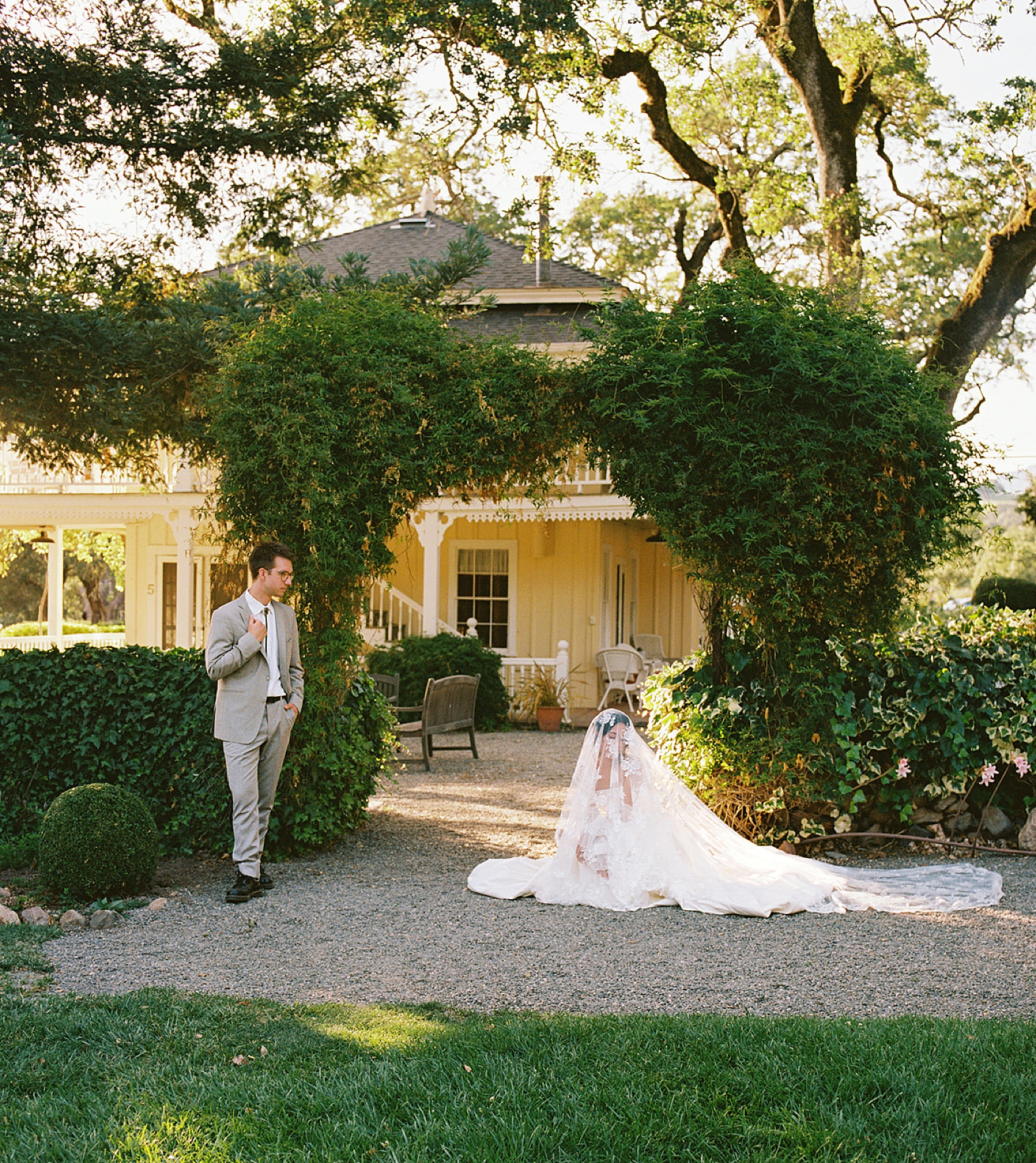 a bride sits on the ground under her veil as her groom looks on in the gardens