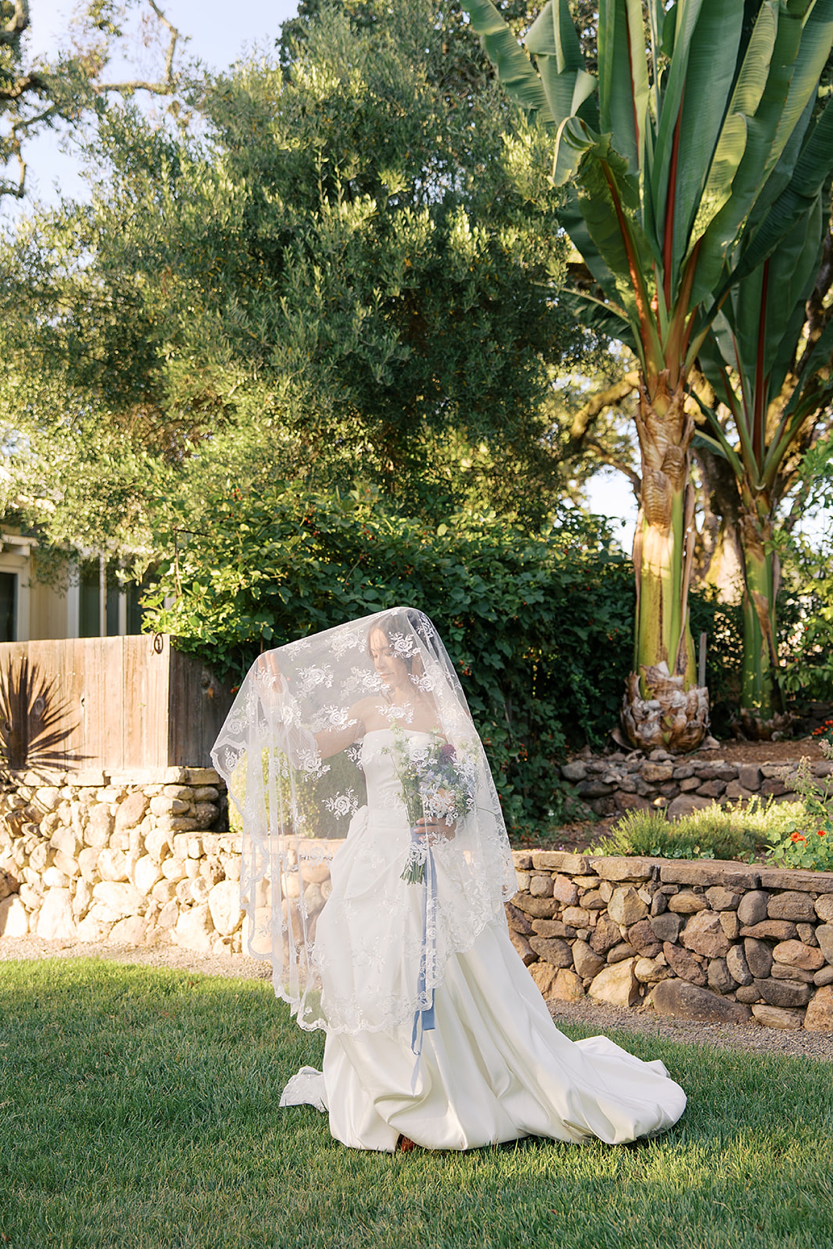 A bride stands in a garden lawn at the beltane ranch wedding venue under her lace embroidered veil