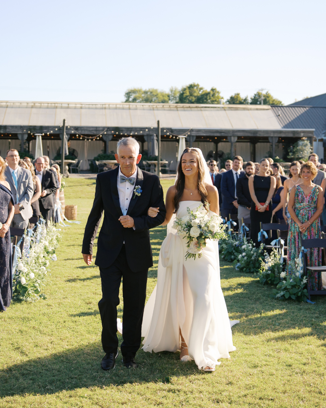 a bride in arms with her dad walking down the aisle smiling looking at her husband