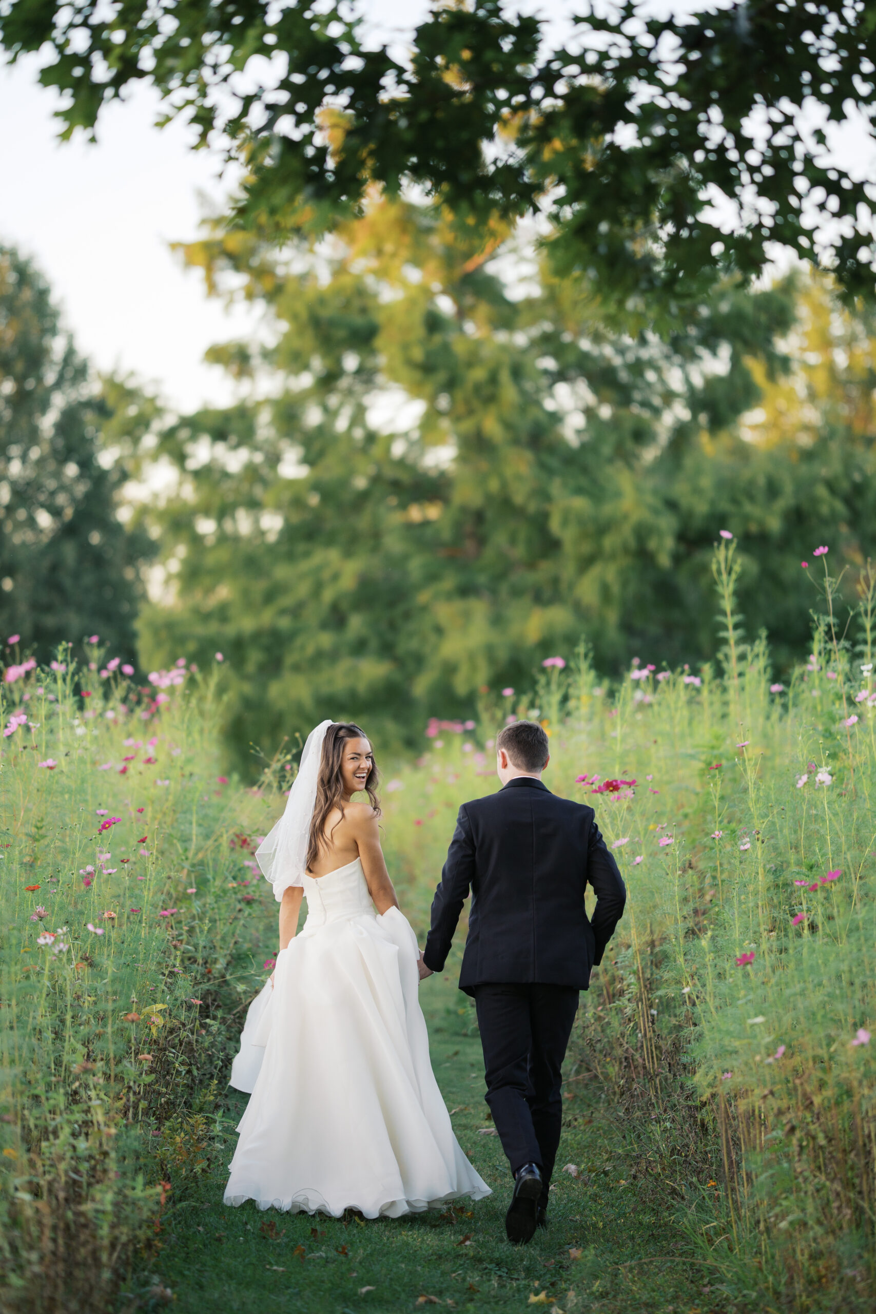 a bride and groom walking away while looking back at the camera in a field of wildflowers at their long hollow gardens wedding