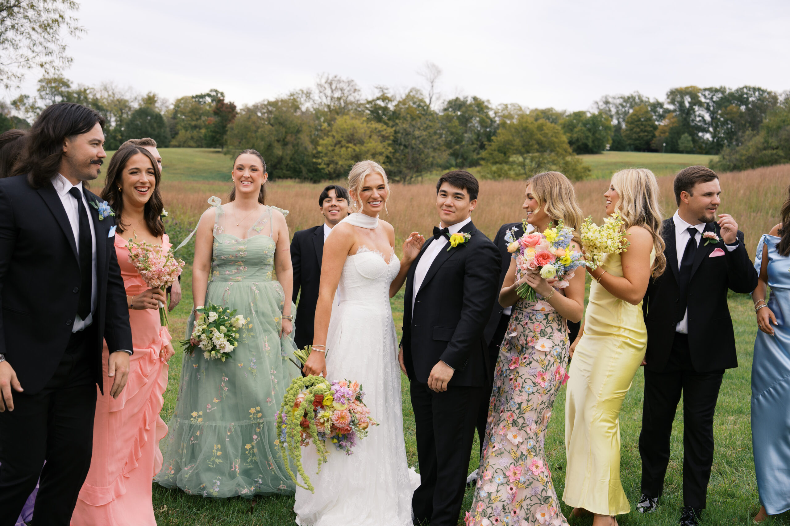 A bridal party dressed in pinks, greens, and yellows surround the bride and groom and are laughing at a mint springs farm wedding.