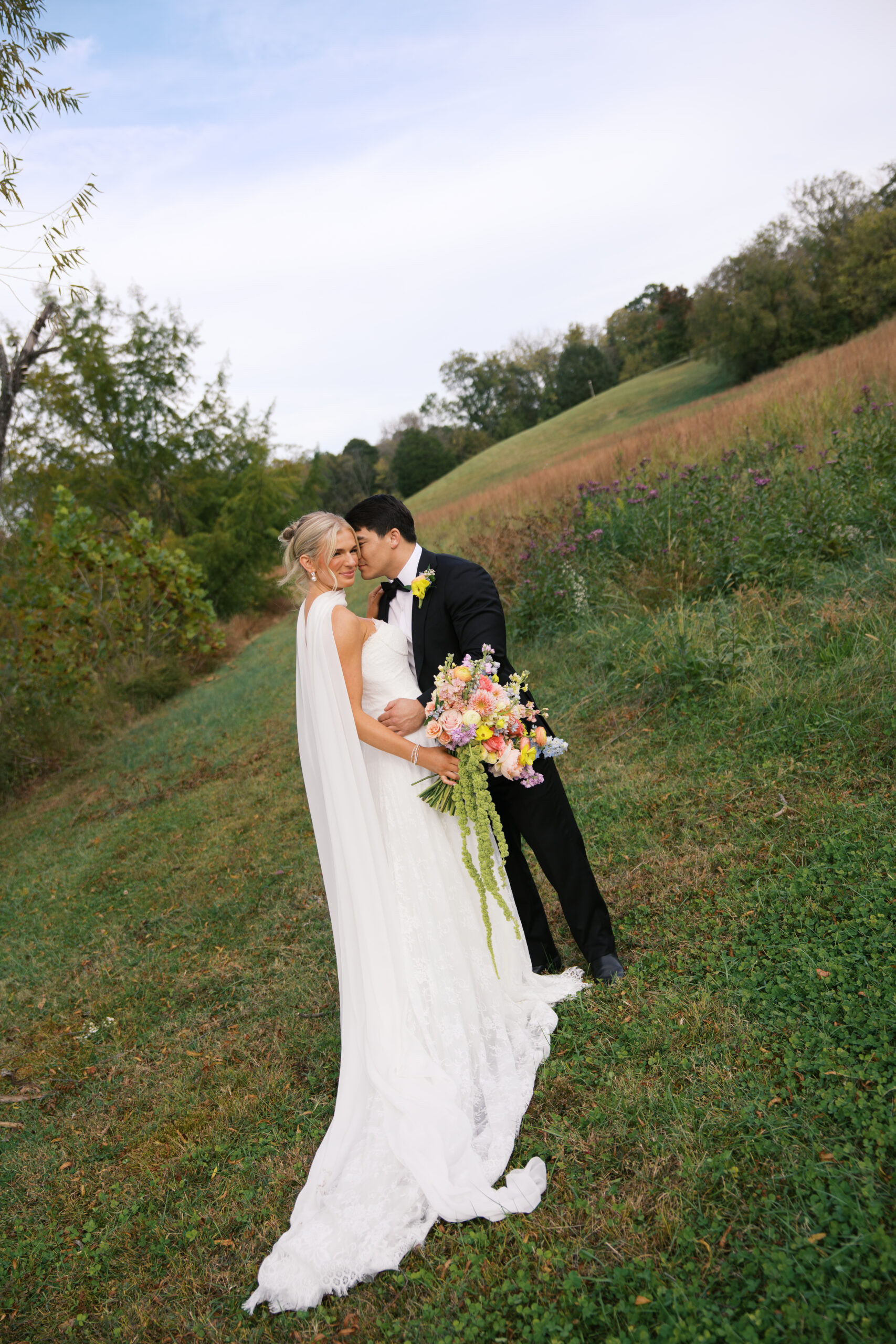 A bride holding a pink and green bouquet next to her groom wearing a black tux kissing her cheek at mint springs farm