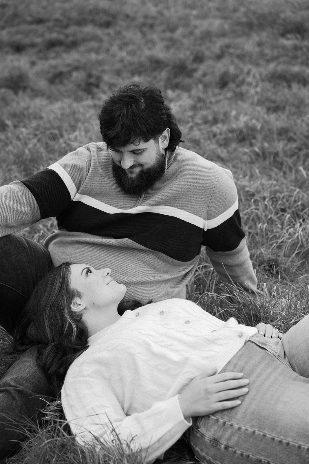 a woman is laying on a mans lap during her engagement photos at harlinsdale farm. hes looking down at her wearing a striped sweater.