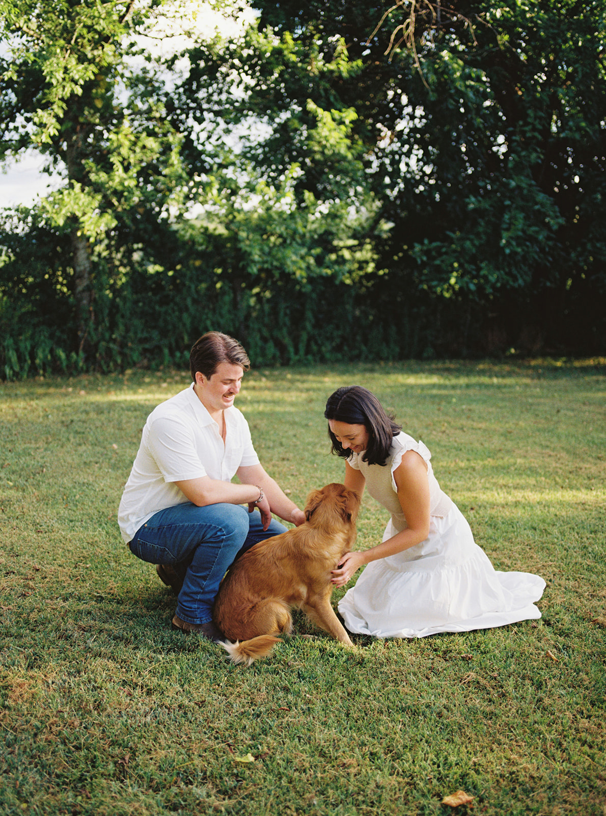 a girl in a white dress kneeling down next to a golden retriever and a man wearing a white shirt and jeans during their engagement photos at bells bend in nashville.
