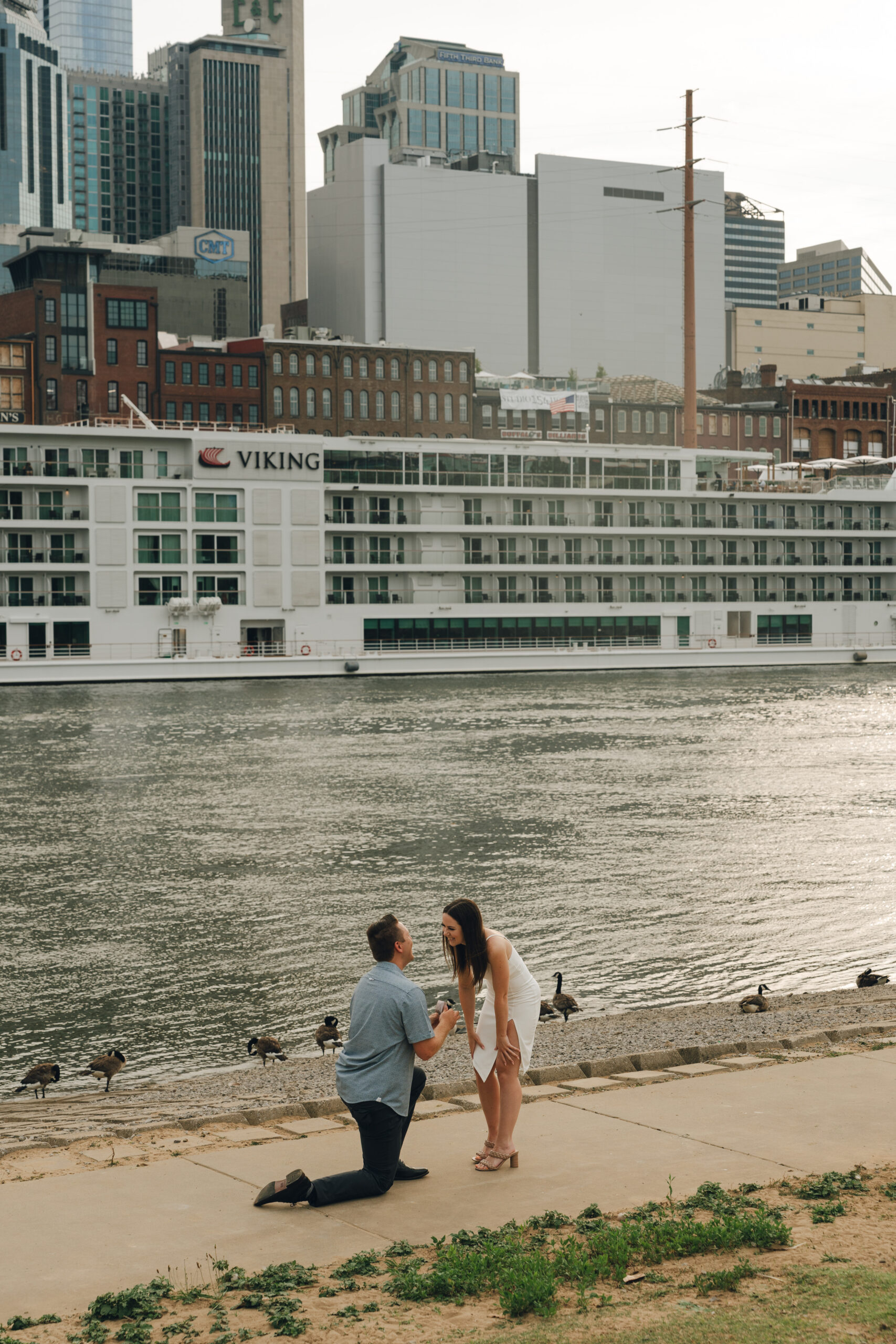 A man on a knee proposing to a woman in a white dress at the cumberland river in nashville.