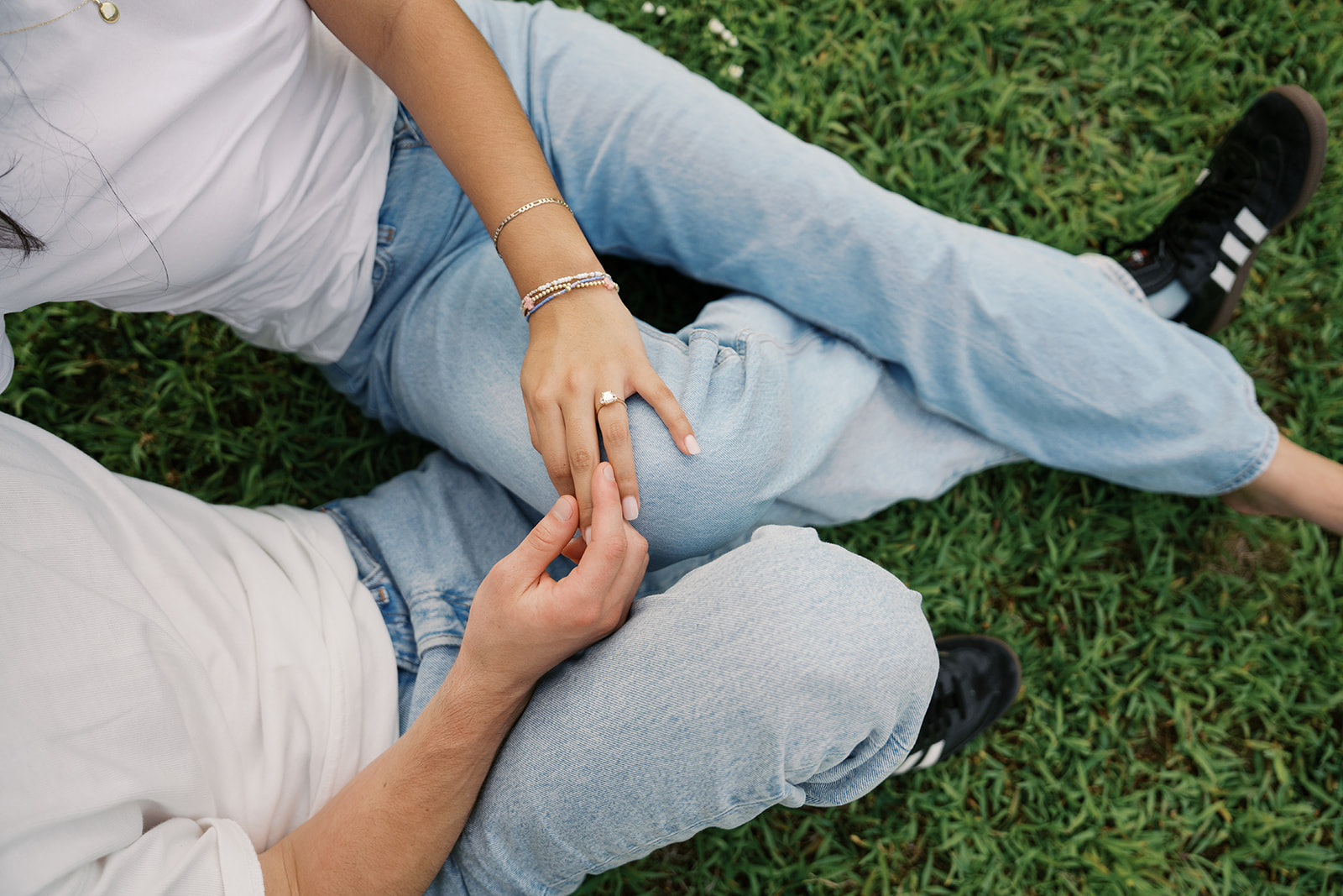 a man and woman are sitting in the grass wearing blue jeans while he plays with her engagement ring