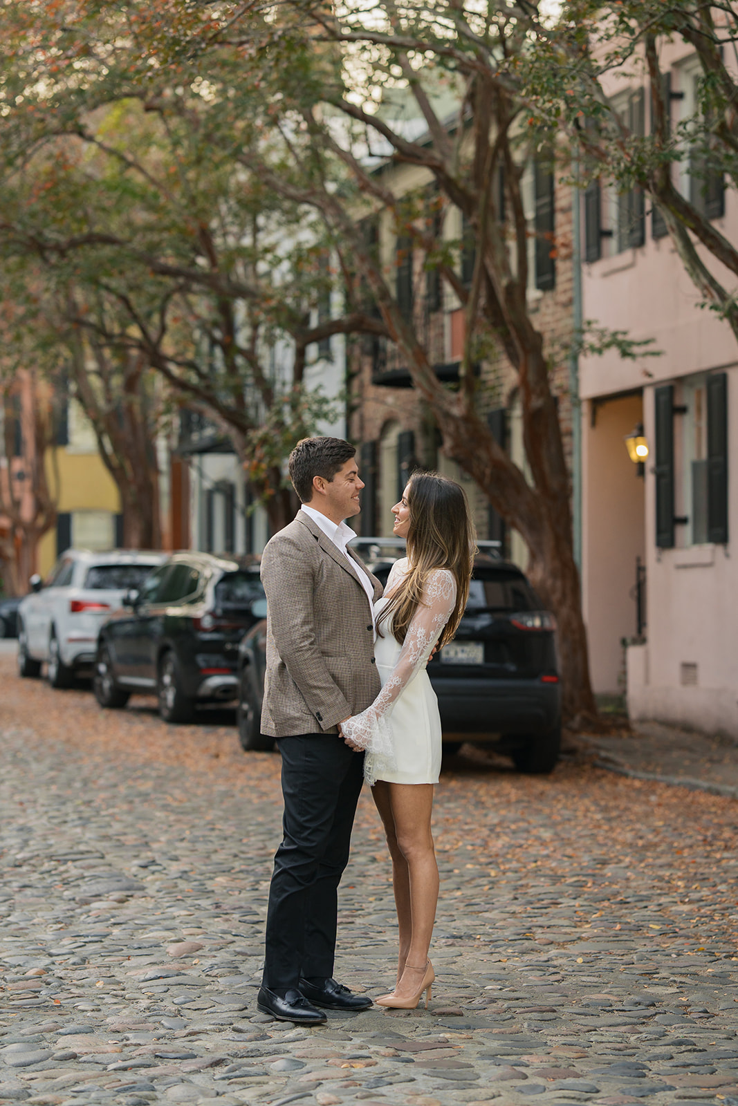 A couple holds hands on a cobblestone street among colorful homes in charleston south carolina during their engagement session