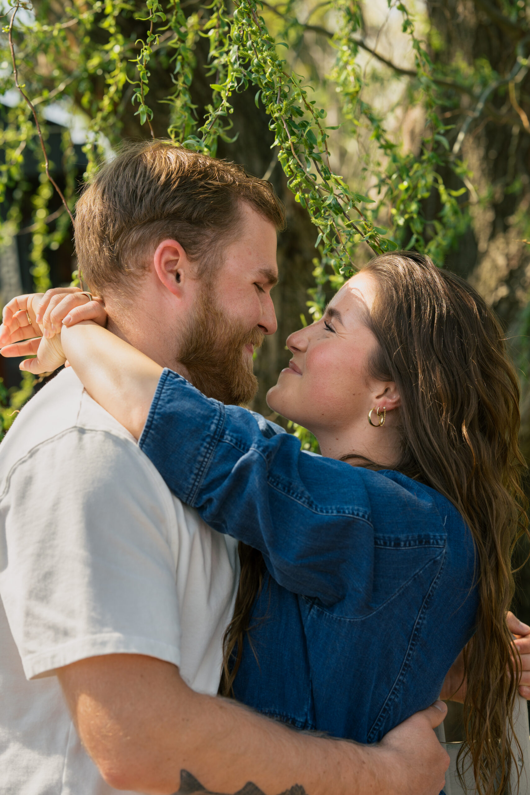 Woman wraps her arms around her fiancés neck as they smile at each other while standing under a tree.