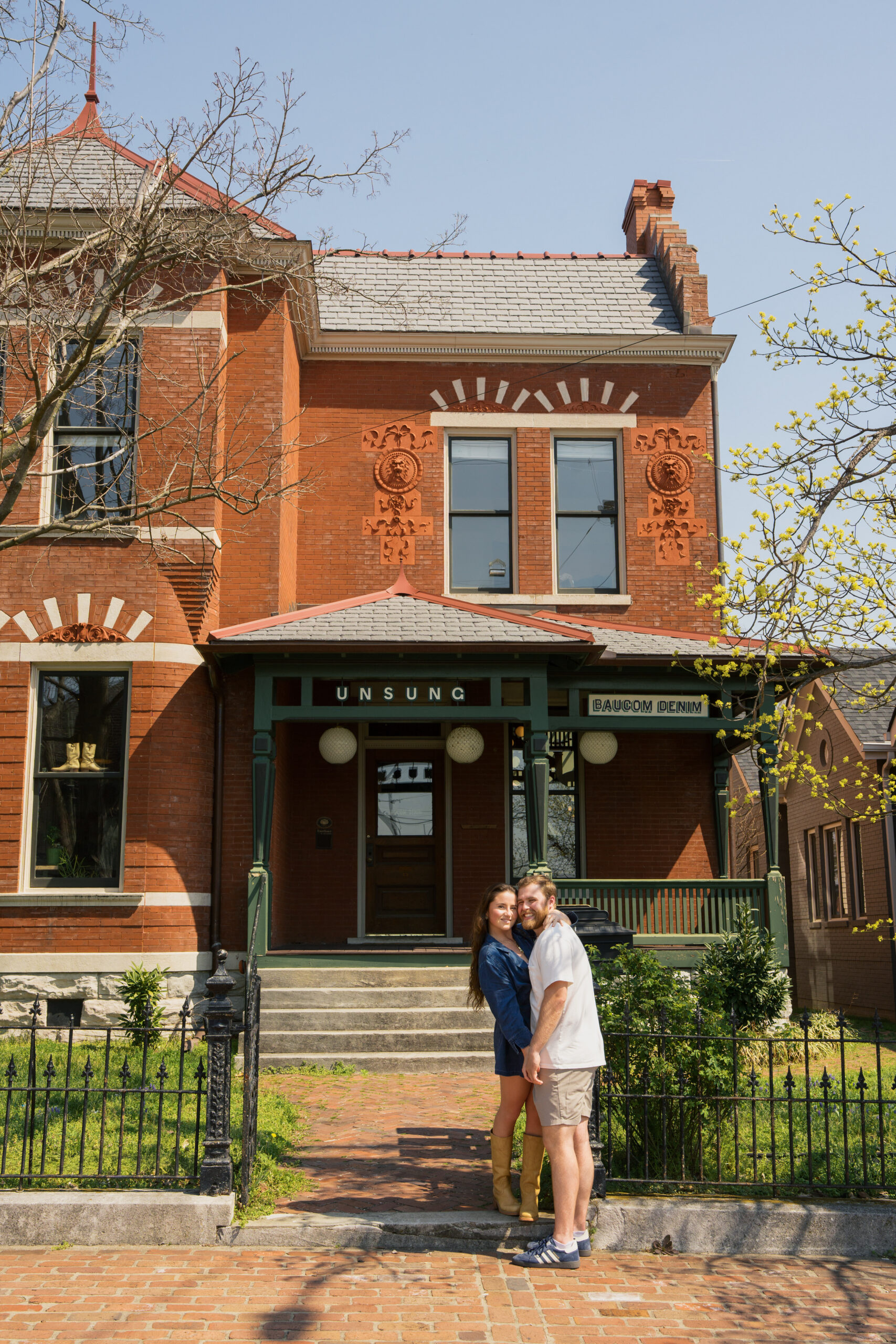 Couples rests sides of their heads together as they smile while standing in front of a colonial style home.