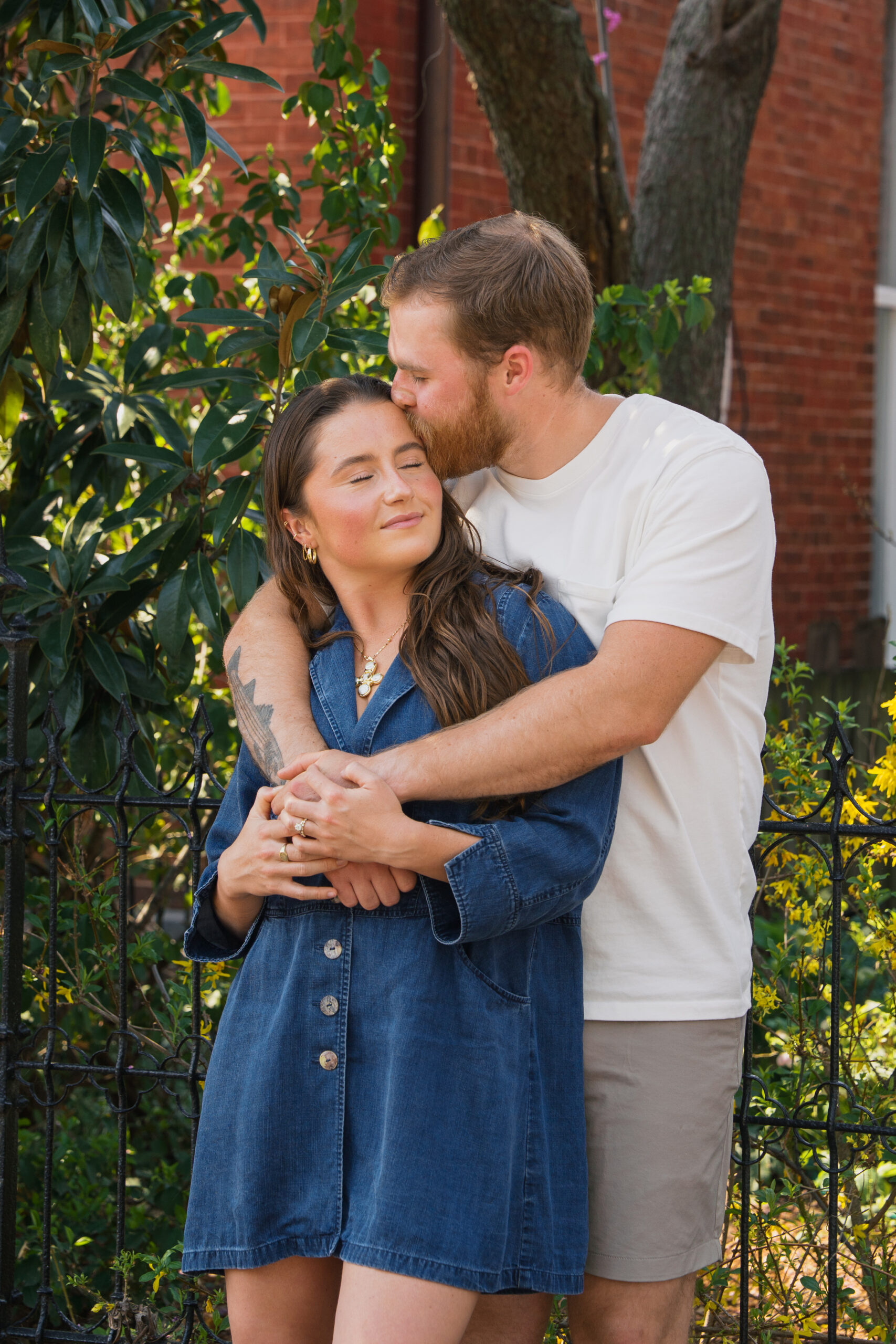 Man wraps his arms around his fiancé as he kisses her forehead and she closes her eyes with a soft smile on her face.