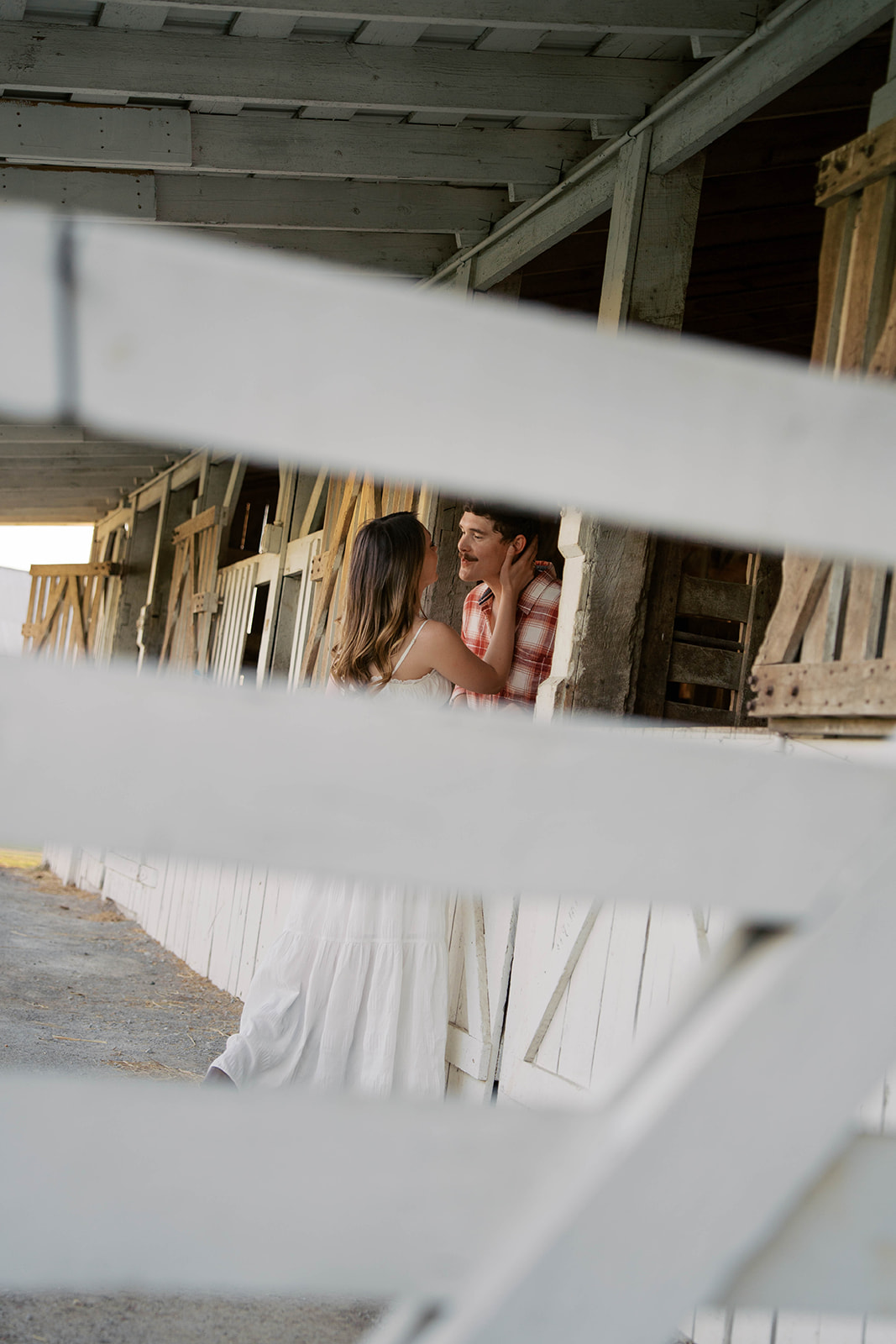 A photo taken from in between wood on a fence of a woman holding the side of her fiancés face while he leans in for a kiss. 