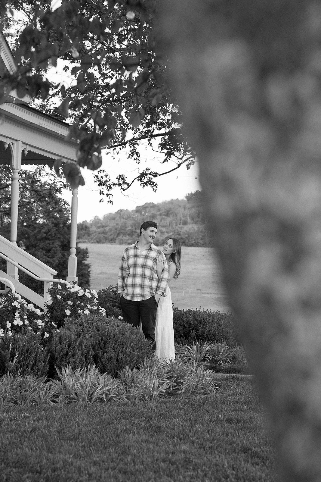 Black and white photo peeking from behind a tree of a woman hugging her fiancé from behind. 