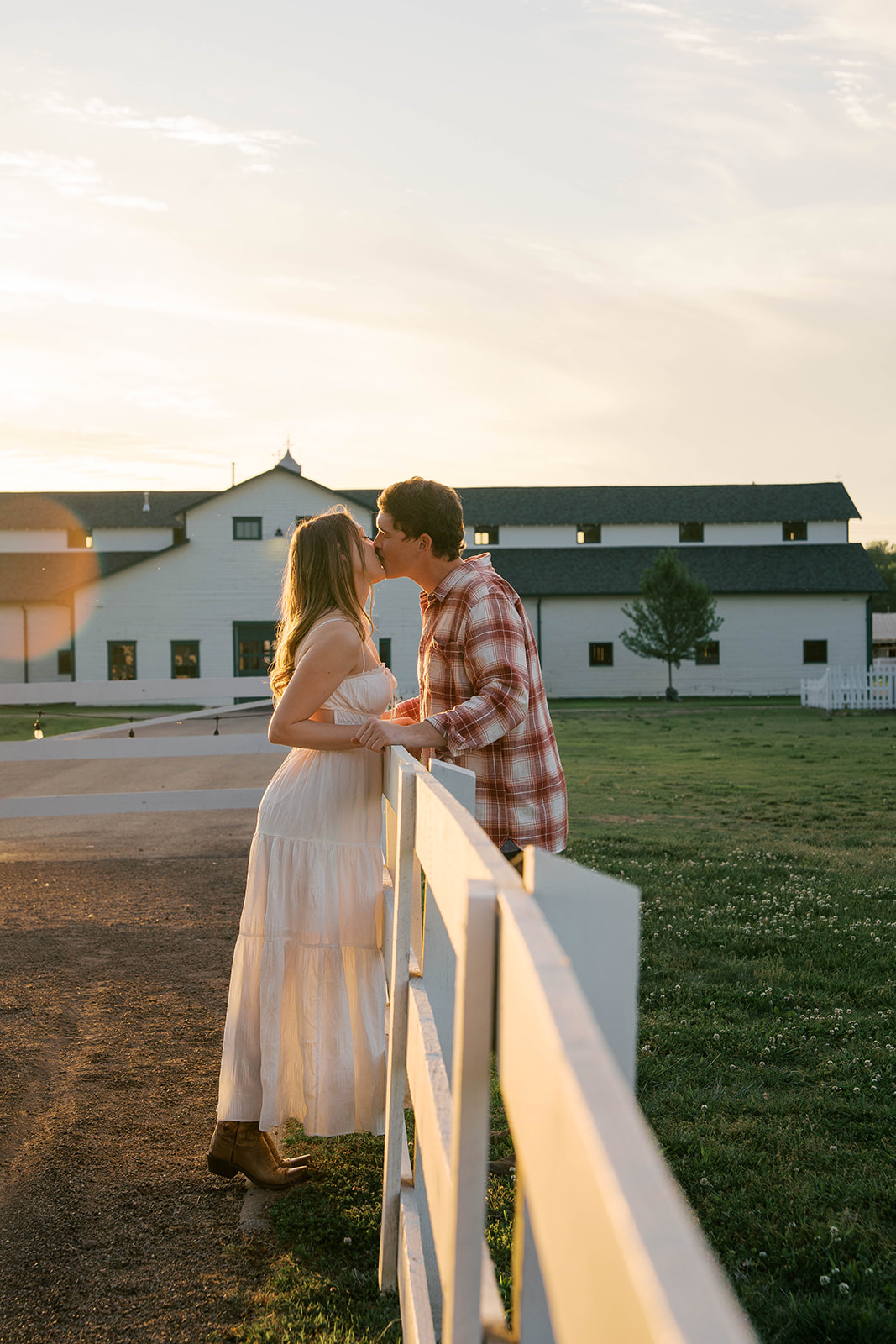 Couple leans over a white fence as they share a kiss in front of a white barn. 
