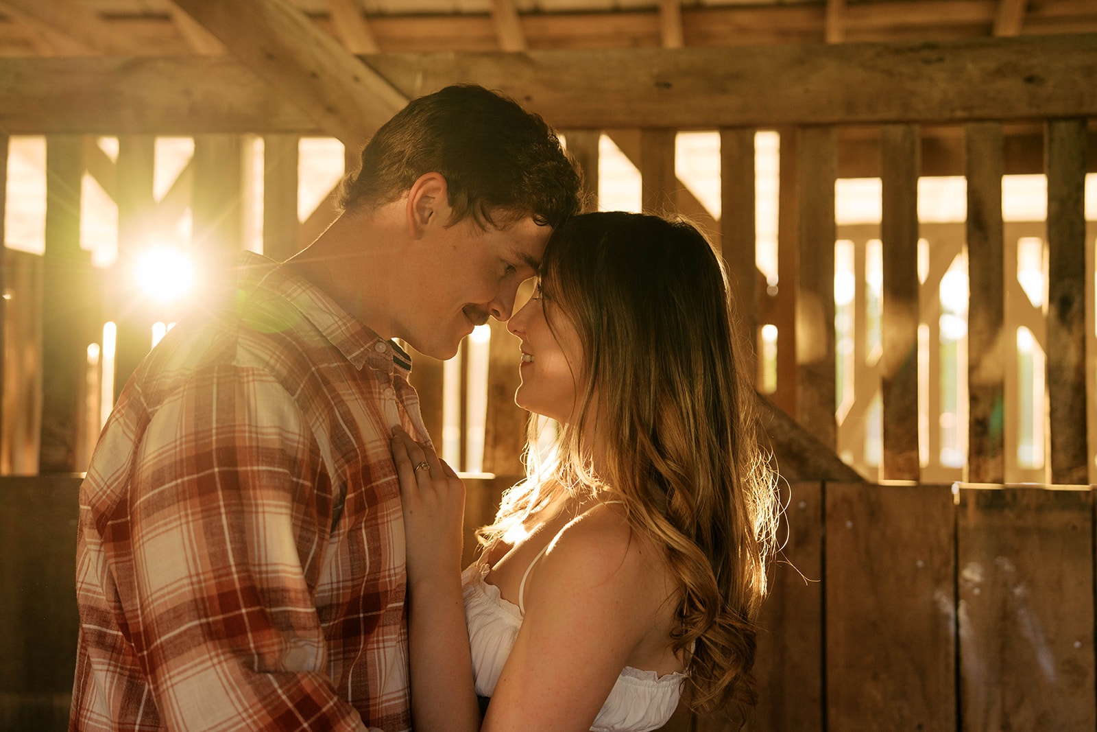 Couple smiles as they rest their foreheads together as the sun shines through the barn doors. 