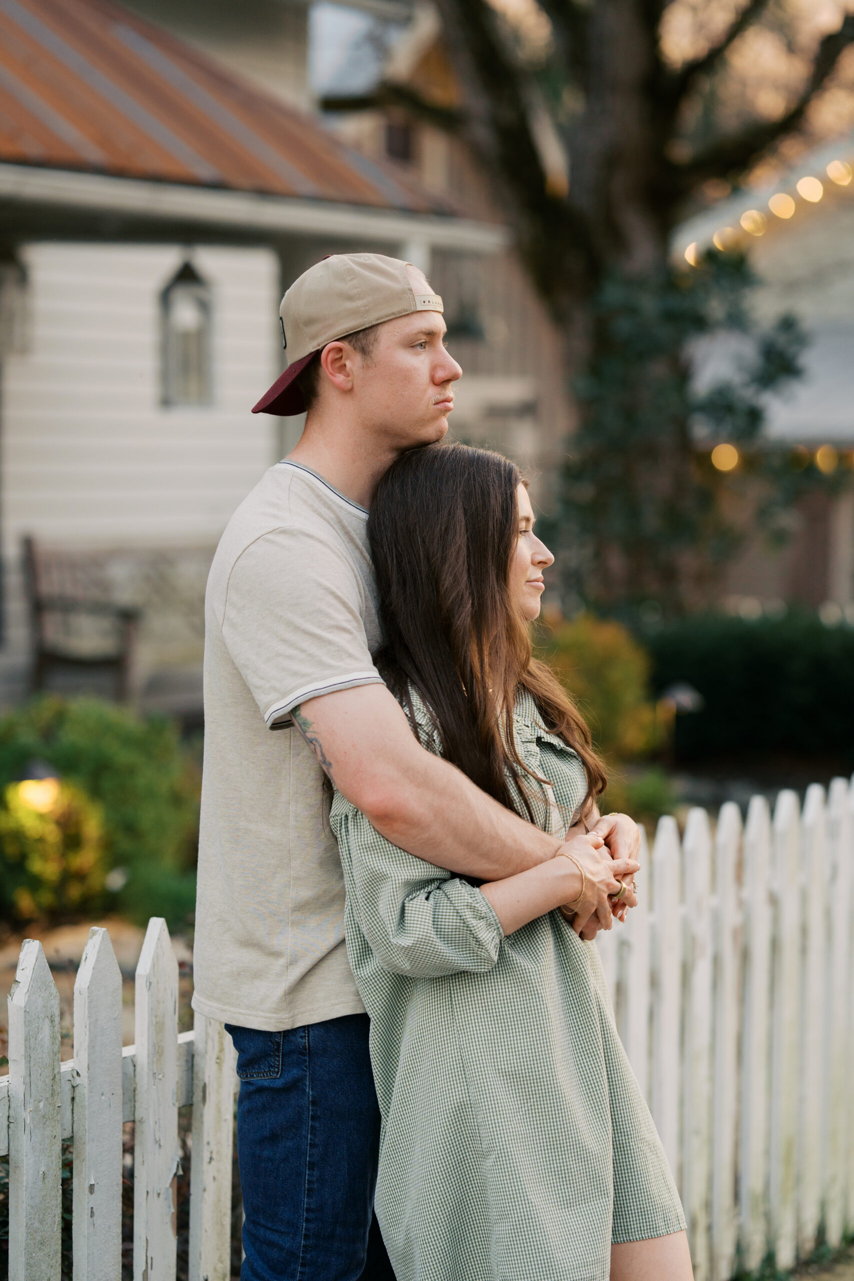 A man stands against a fence with his hat on backwards holding his wife wearing a green dress during their couples session in leipers fork, tennessee.