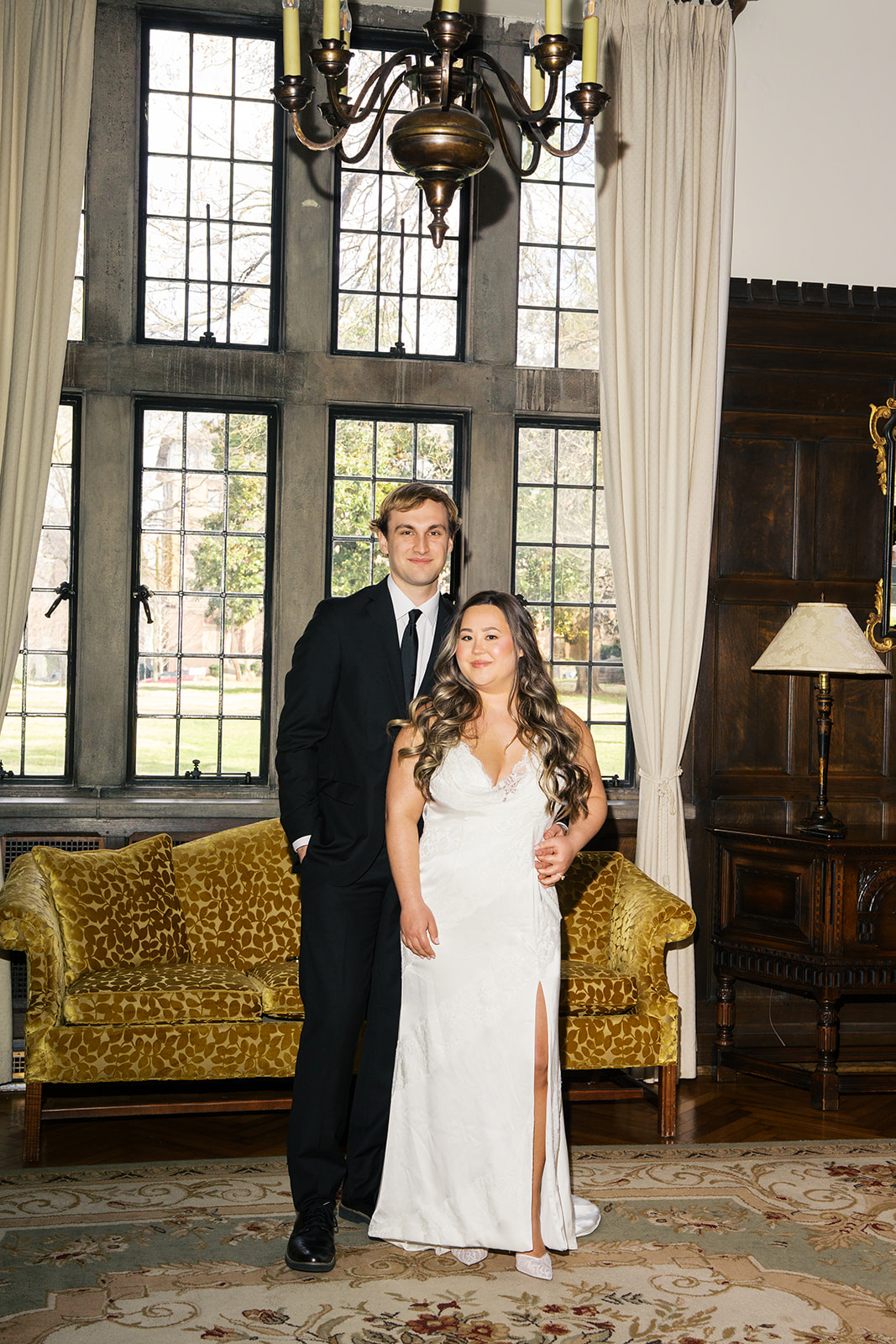 Couple dressed in formal attire stands in front of a big window and they give a soft smile to the camera.