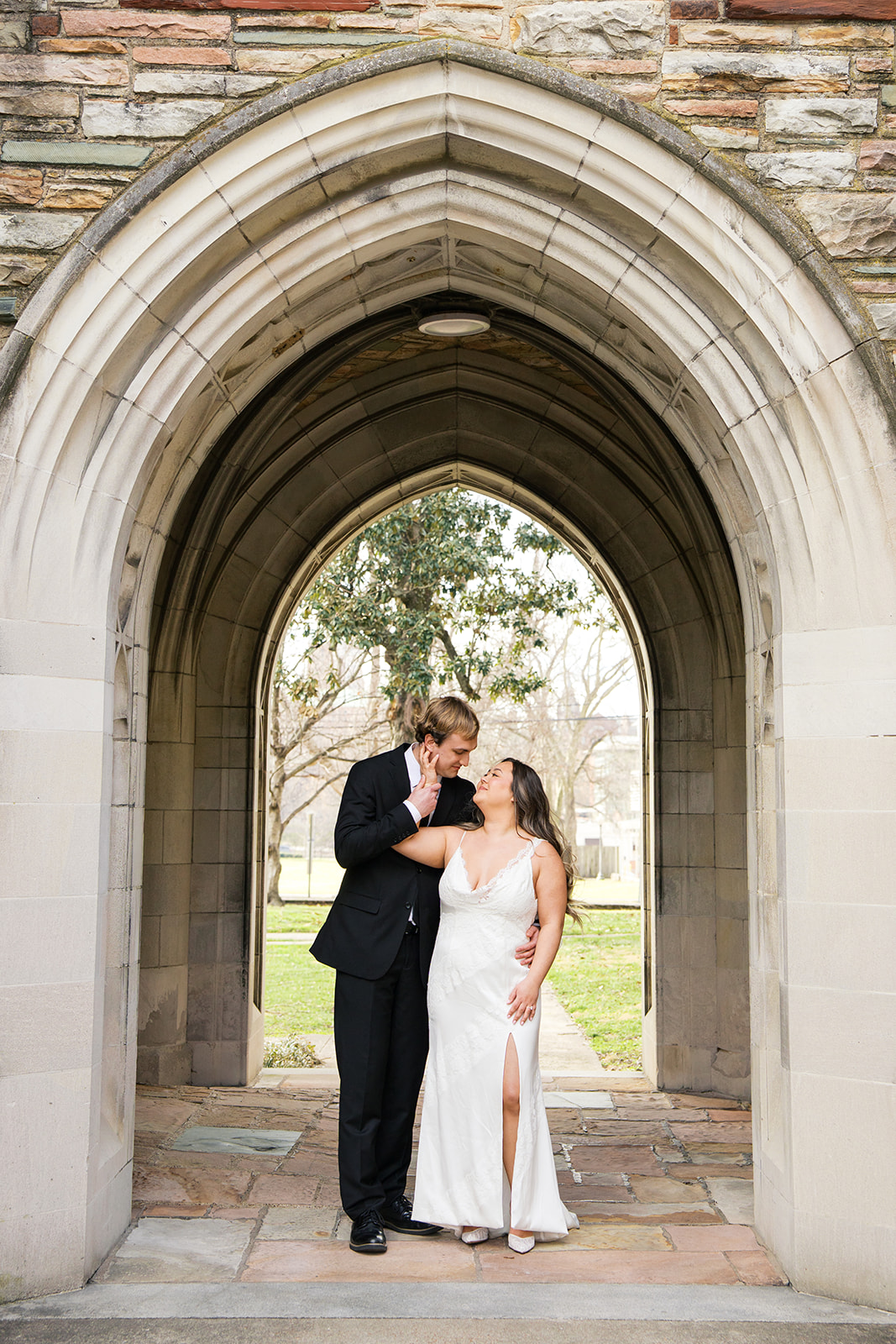 A bride reaches her hand up to hold the grooms face behind her under an arch at scarritt bennett center during their wedding portraits