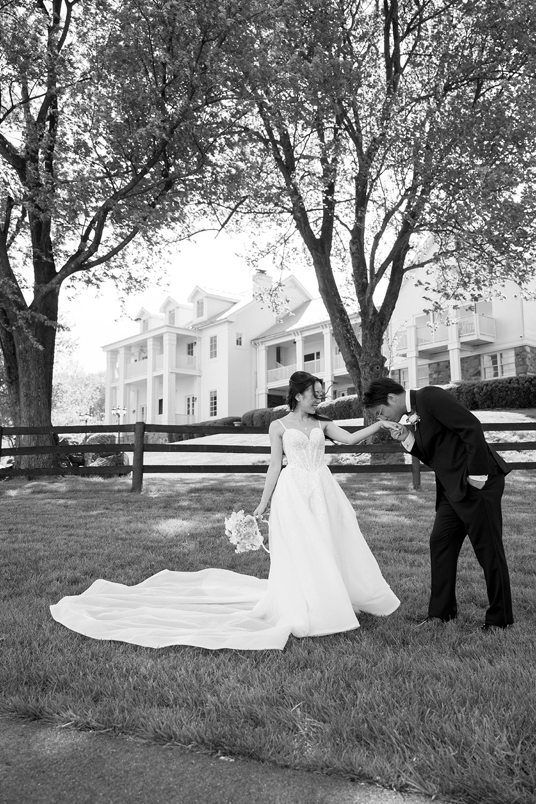 A groom kissing the hand of a bride in black and white with the view at bluemont wedding venue behind them