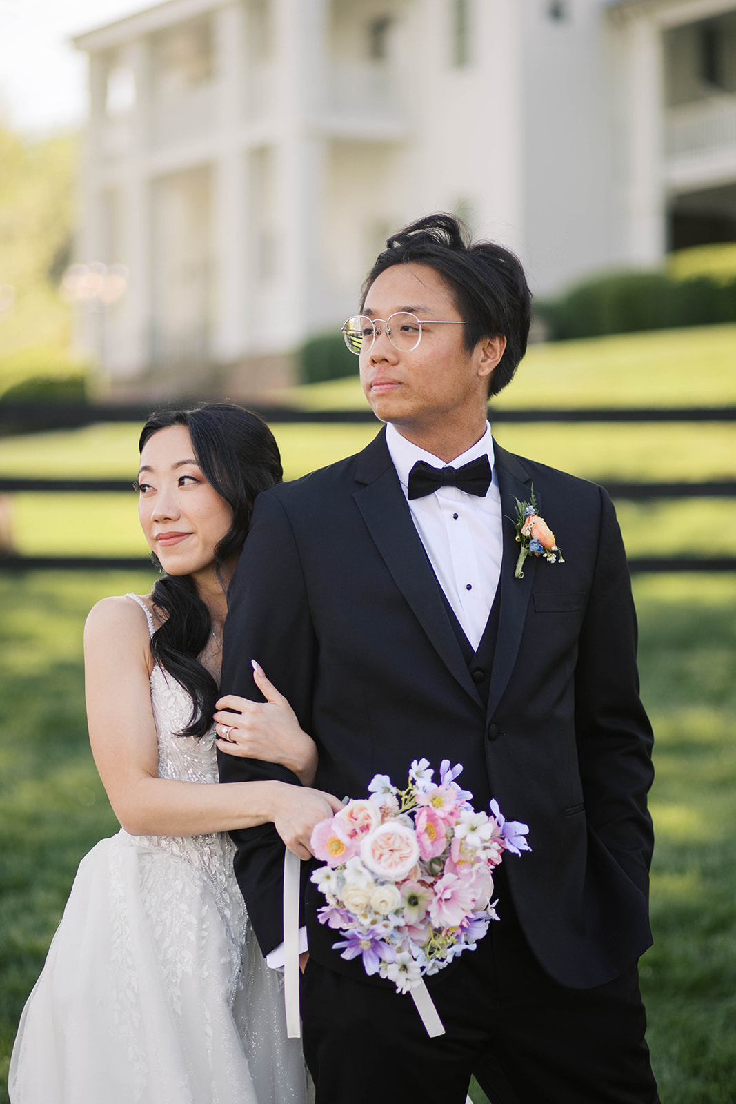 A bride wearing a white floral dress holding onto a groom wearing a black tux looking away from the camera with the view at bluemont venue in the background.