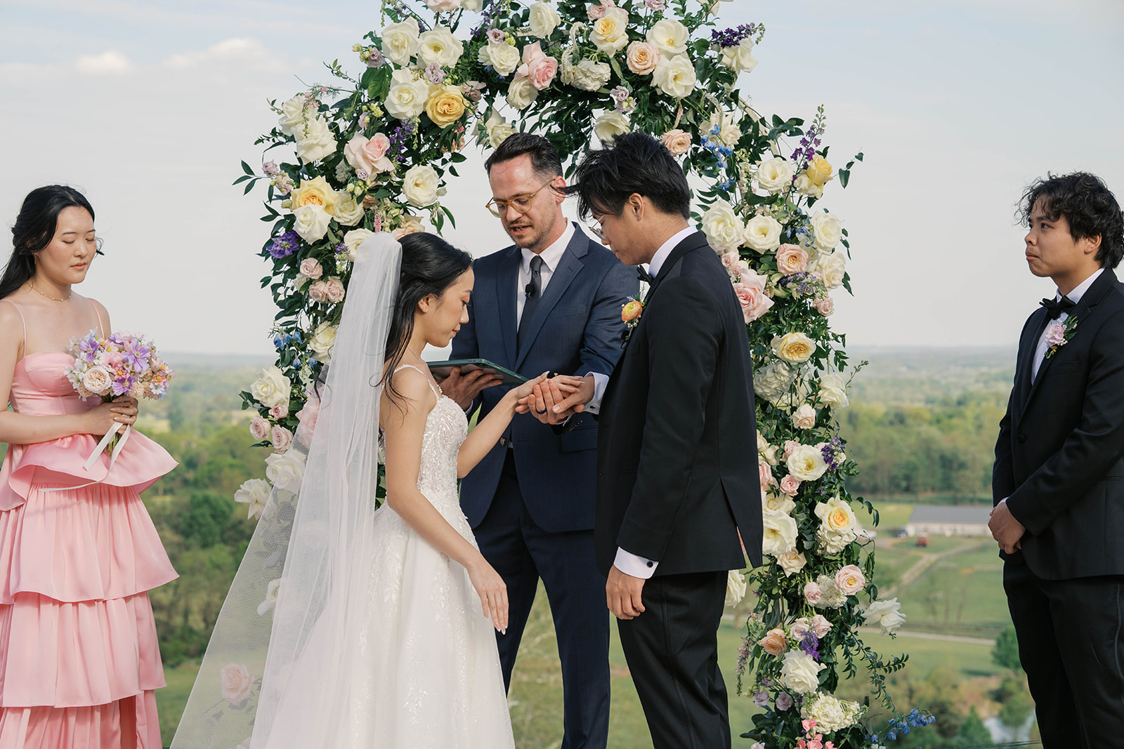 The ring exchange during a wedding at the view at bluemont in virginia with a large yellow and white floral arch behind the couple.