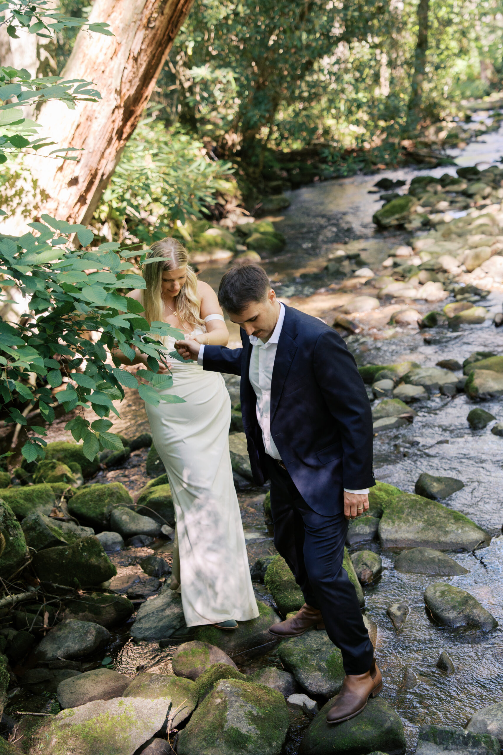 A groom holding hands with a bride leading her through a creek during their elopement in tennessee