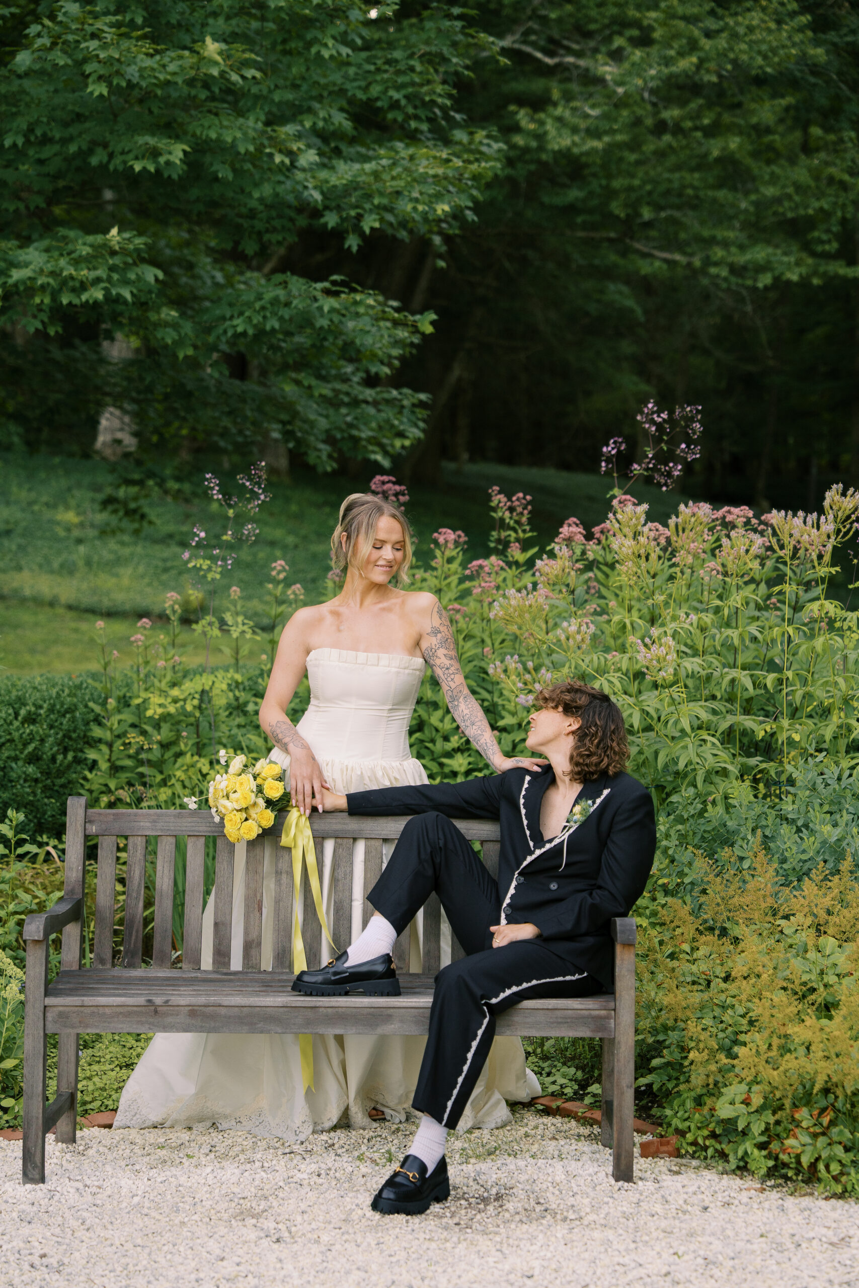 Two brides during their wedding portraits. One is sitting on a bench with their leg up, the other is behind the bench holding the others shoulder. They look at each other and are smiling with flowers in the background.