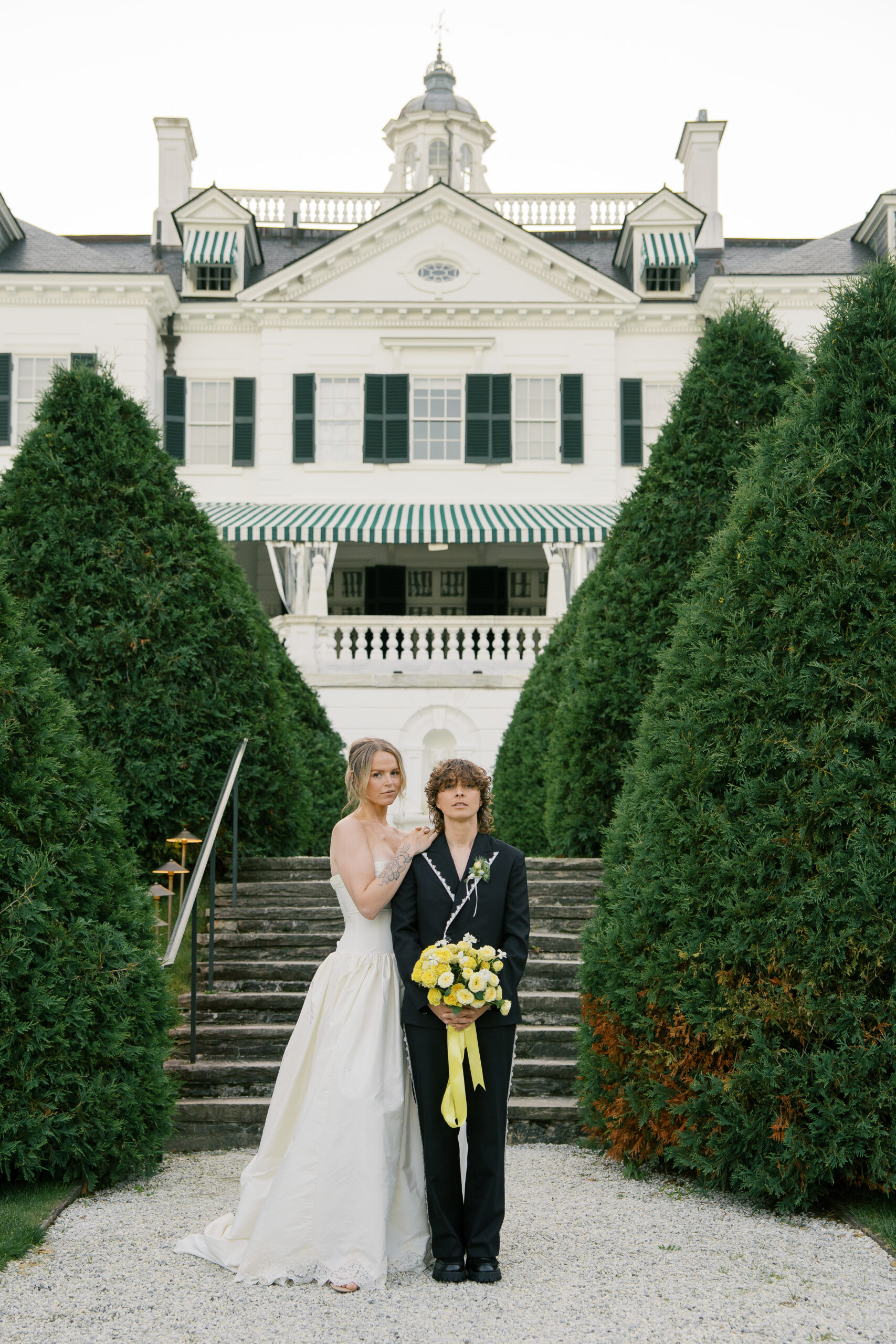 Two brides, one wearing a white wedding dress and one wearing a black lace suit, face the camera smiling during their wedding at the mount in Lenox massachusetts.