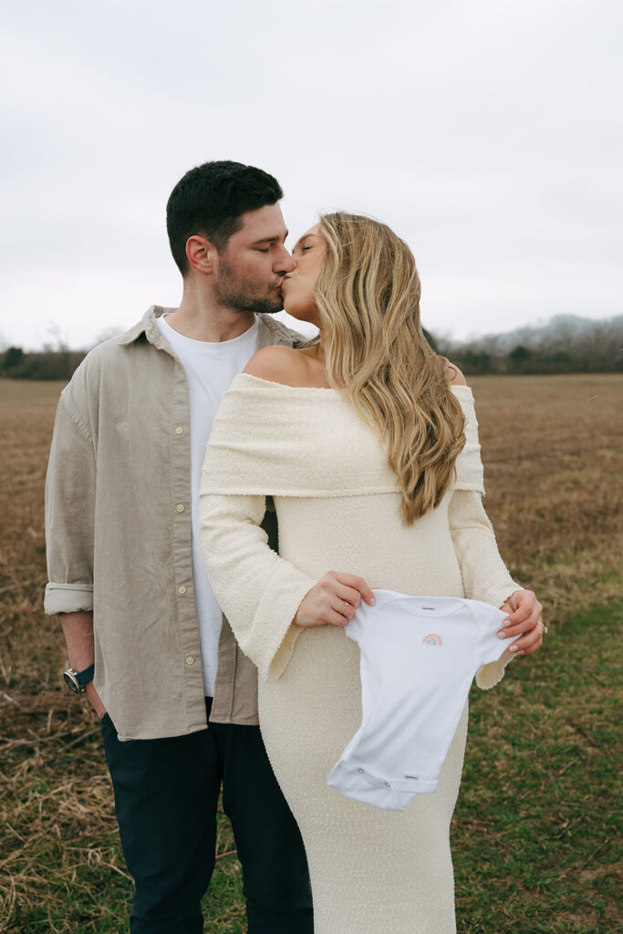 pregnant couple kissing and holding baby onesie 