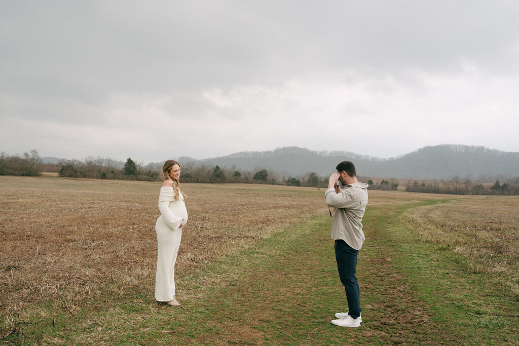 man taking picture of pregnant woman in a white dress at a field in nashville 