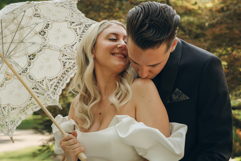 groom kissing bride on the shoulder while holding a parasol